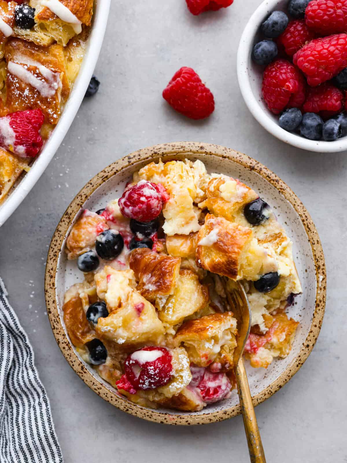 If you love sweet breakfasts, you're in for a treat. This croissant breakfast casserole features tender croissant pieces and fresh berries all coated in a cream cheese custard. As if that's not good enough, everything is topped off with a homemade lemon glaze that's absolutely heavenly. It's love at first bite! A scoop of the casserole served in a stoneware bowl.