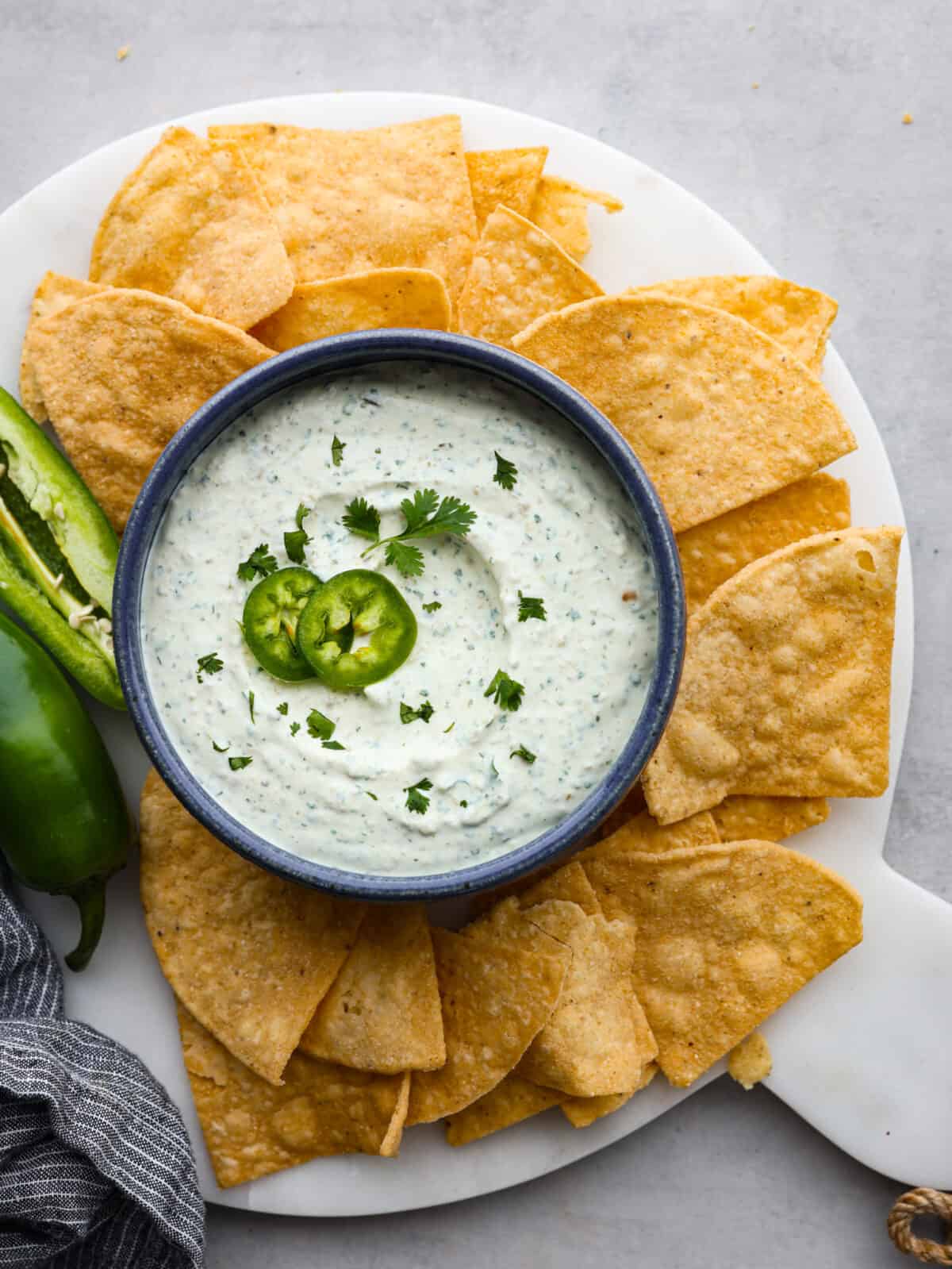 This zesty Jalapeño Dip is the ultimate crowd-pleaser! Blending cream cheese, sour cream, and canned pickled jalapenos with seasoning, it delivers a delightful mix of creaminess and heat. The top view of a platter of tortilla chips with a bowl of jalapeño dip with a sliced jalapeño garnishing the dip.