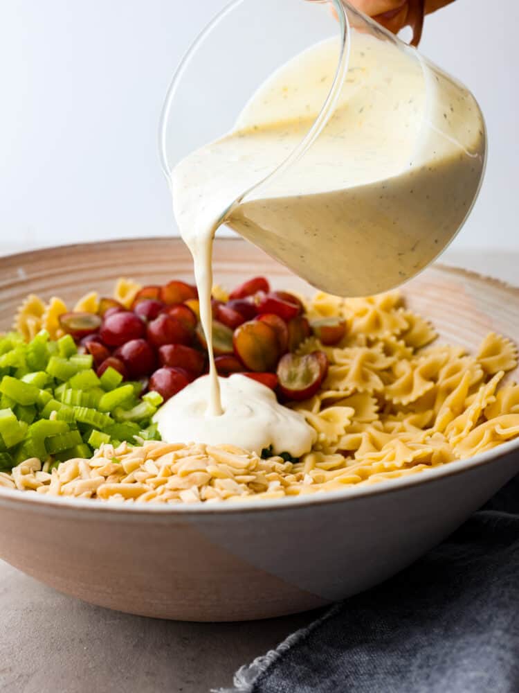 This Lemon Tarragon Pasta Salad combines tender bowtie pasta with crisp celery, juicy red grapes, and slivered almonds all tossed in a creamy lemon yogurt dressing. This refreshing salad complements any dish and is perfect for even the pickiest of eaters! Dressing being poured over pasta salad ingredients.
