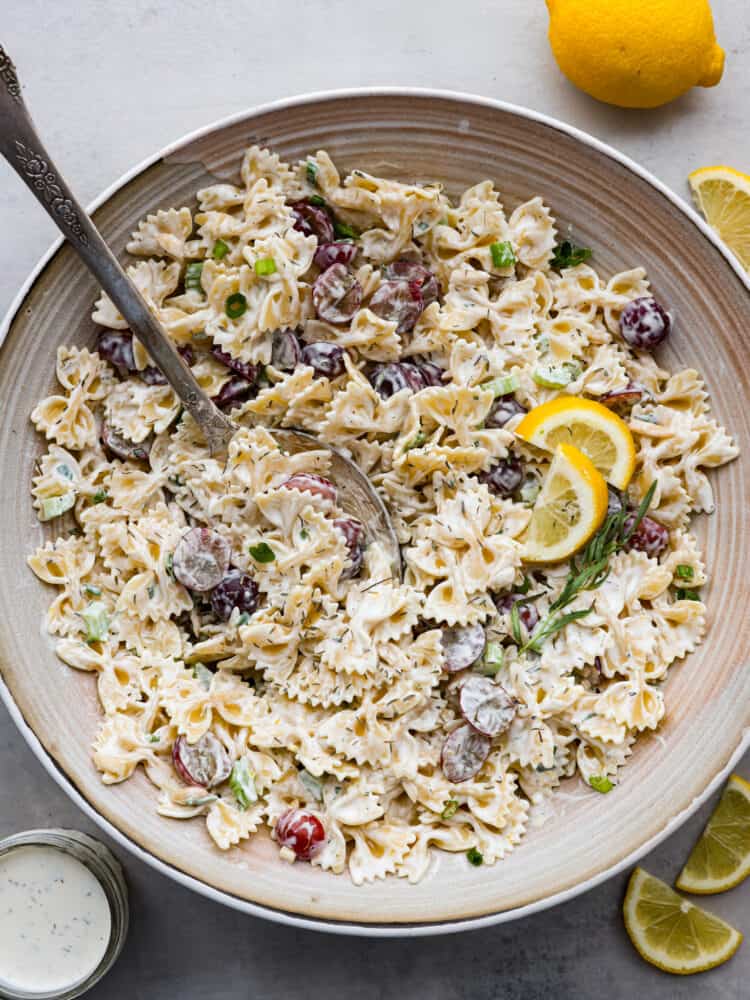 This Lemon Tarragon Pasta Salad combines tender bowtie pasta with crisp celery, juicy red grapes, and slivered almonds all tossed in a creamy lemon yogurt dressing. This refreshing salad complements any dish and is perfect for even the pickiest of eaters! The top view of a large bowl with a silver spoon that has lemon tarragon pasta salad in it.