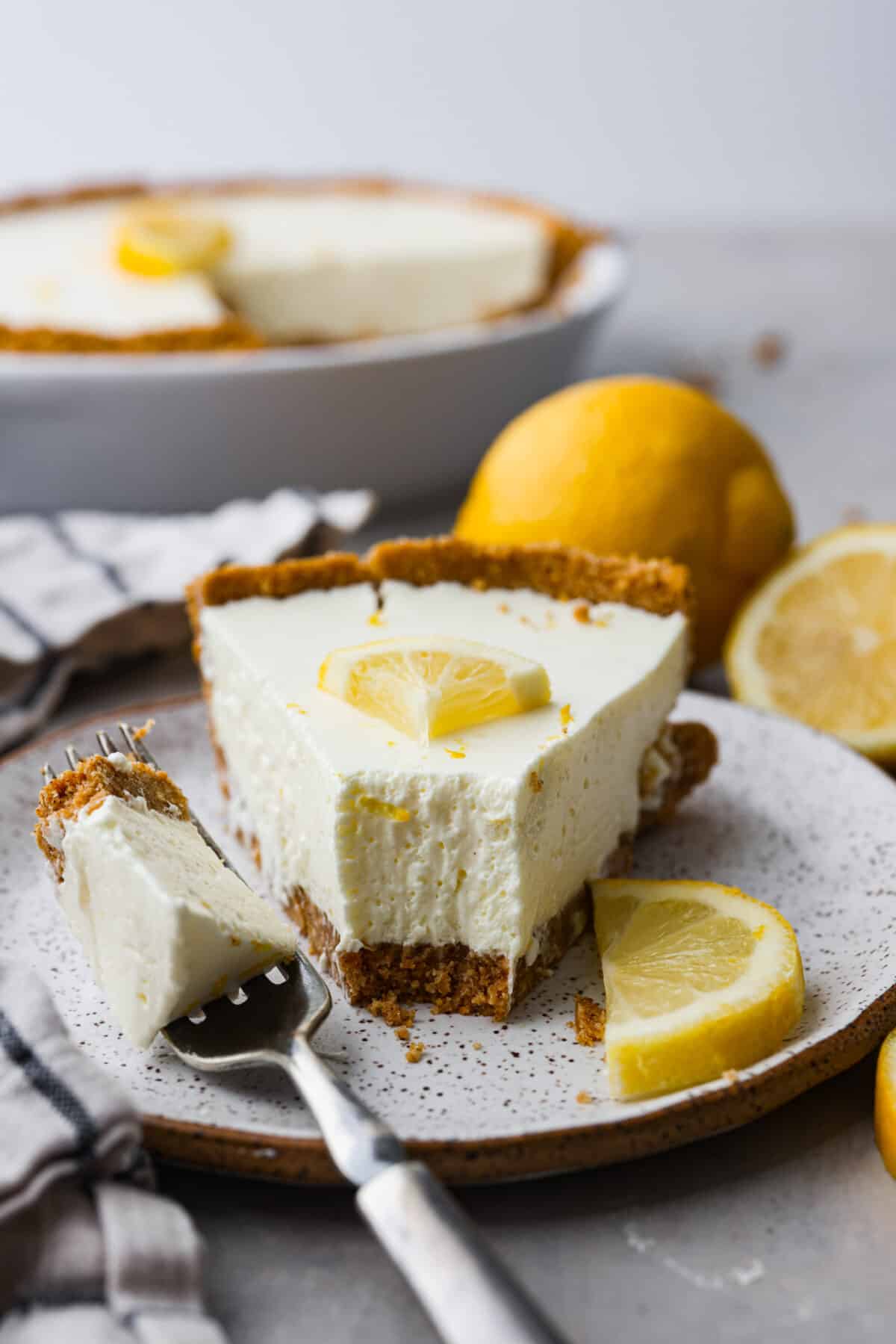 No-bake lemon pie is a light and creamy pie bursting with refreshing lemony flavor. It's sweet and zingy all at the same time! This pie is easy to make and is the perfect spring or summer dessert! Close view of no-bake lemon pie on a white plate. Garnished with lemon slices and a fork on the plate.