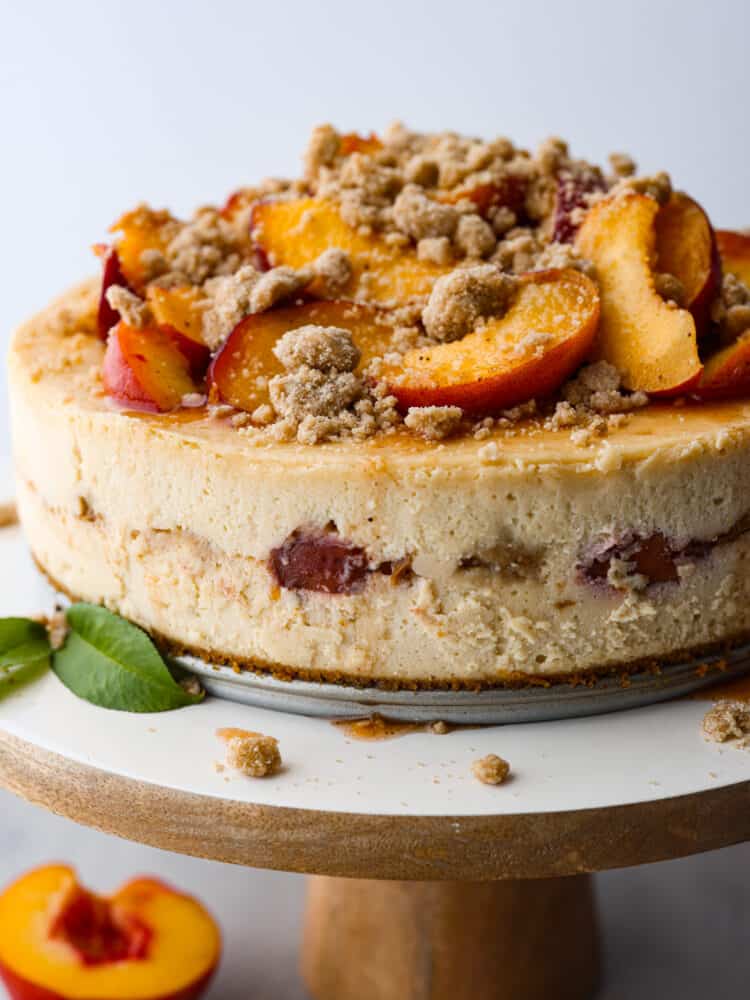 Indulge in a delightful treat with this mouthwatering Peach Cobbler Cheesecake recipe. A delicious combination of creamy cheesecake, caramelized peaches, and a buttery streusel crust, all perfectly balanced to create a dessert that will impress any crowd! A peach cobbler cheesecake on a wooden and white cake stand.