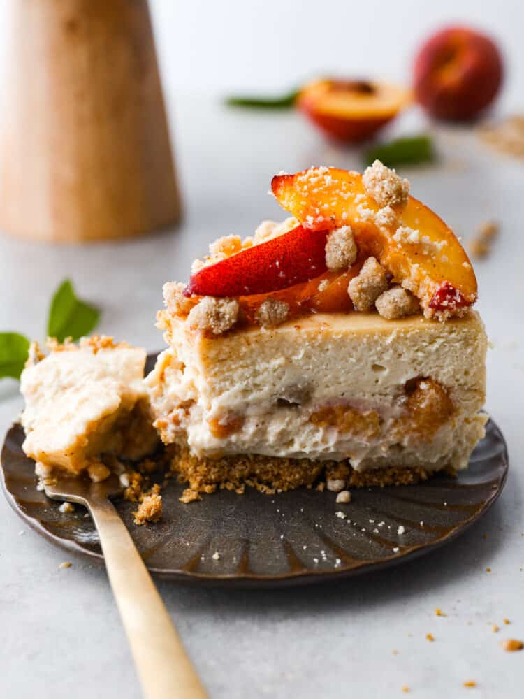Indulge in a delightful treat with this mouthwatering Peach Cobbler Cheesecake recipe. A delicious combination of creamy cheesecake, caramelized peaches, and a buttery streusel crust, all perfectly balanced to create a dessert that will impress any crowd! A slice of peach cobbler cheesecake on a brown plate with a gold fork.