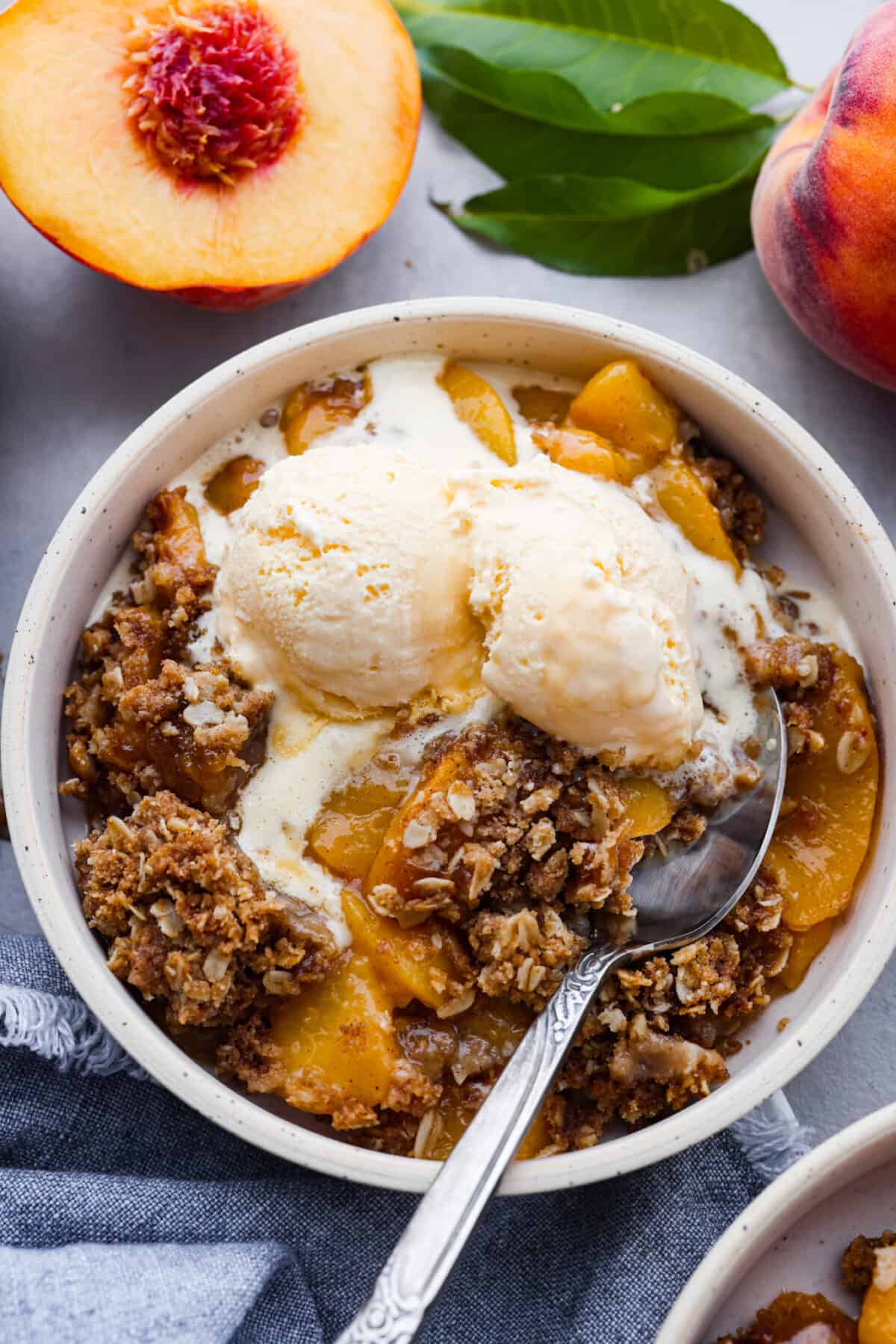 Celebrate summer with a mouthwatering Peach Crisp! Juicy peaches, sweet cinnamon, and a crunchy topping come together for a delightful treat that's perfect when it's served warm with a scoop of vanilla ice cream! Peach crisp in a bowl with a silver spoon and a scoop of ice cream.