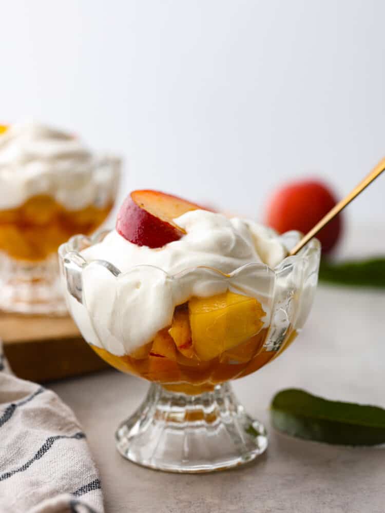 Peaches and cream is a simple and refreshing summertime dessert. Ripe peaches lightly sweetened with sugar and vanilla bean paste, then topped with fresh homemade whipped cream. Every summer we make this classic treat that is bursting with sweet peaches and fluffy cream! Close view of peaches and cream in a glass dish with a gold spoon. A striped towel and peach leaves are next to the dish.