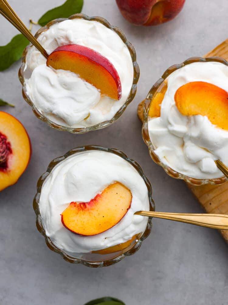 Peaches and cream is a simple and refreshing summertime dessert. Ripe peaches lightly sweetened with sugar and vanilla bean paste, then topped with fresh homemade whipped cream. Every summer we make this classic treat that is bursting with sweet peaches and fluffy cream! Top view of three glass dishes filled with peaches and cream. Gold spoons are nestled inside the dishes.
