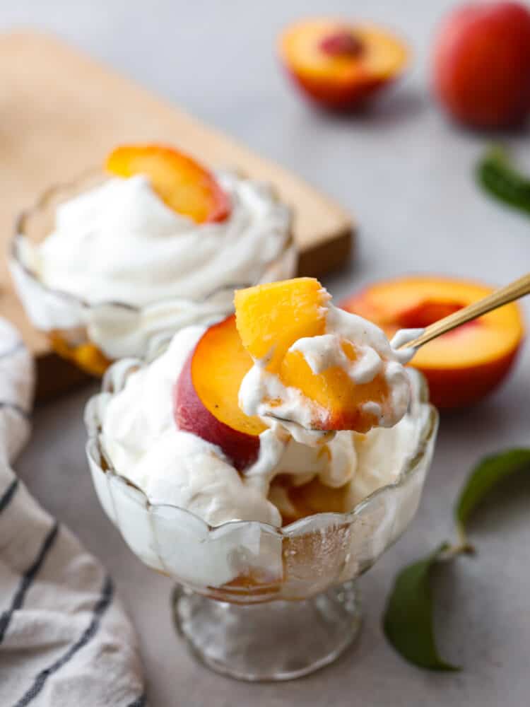 Peaches and cream is a simple and refreshing summertime dessert. Ripe peaches lightly sweetened with sugar and vanilla bean paste, then topped with fresh homemade whipped cream. Every summer we make this classic treat that is bursting with sweet peaches and fluffy cream! Close view of peaches and cream with a gold spoon lifting up peaches from the dish.