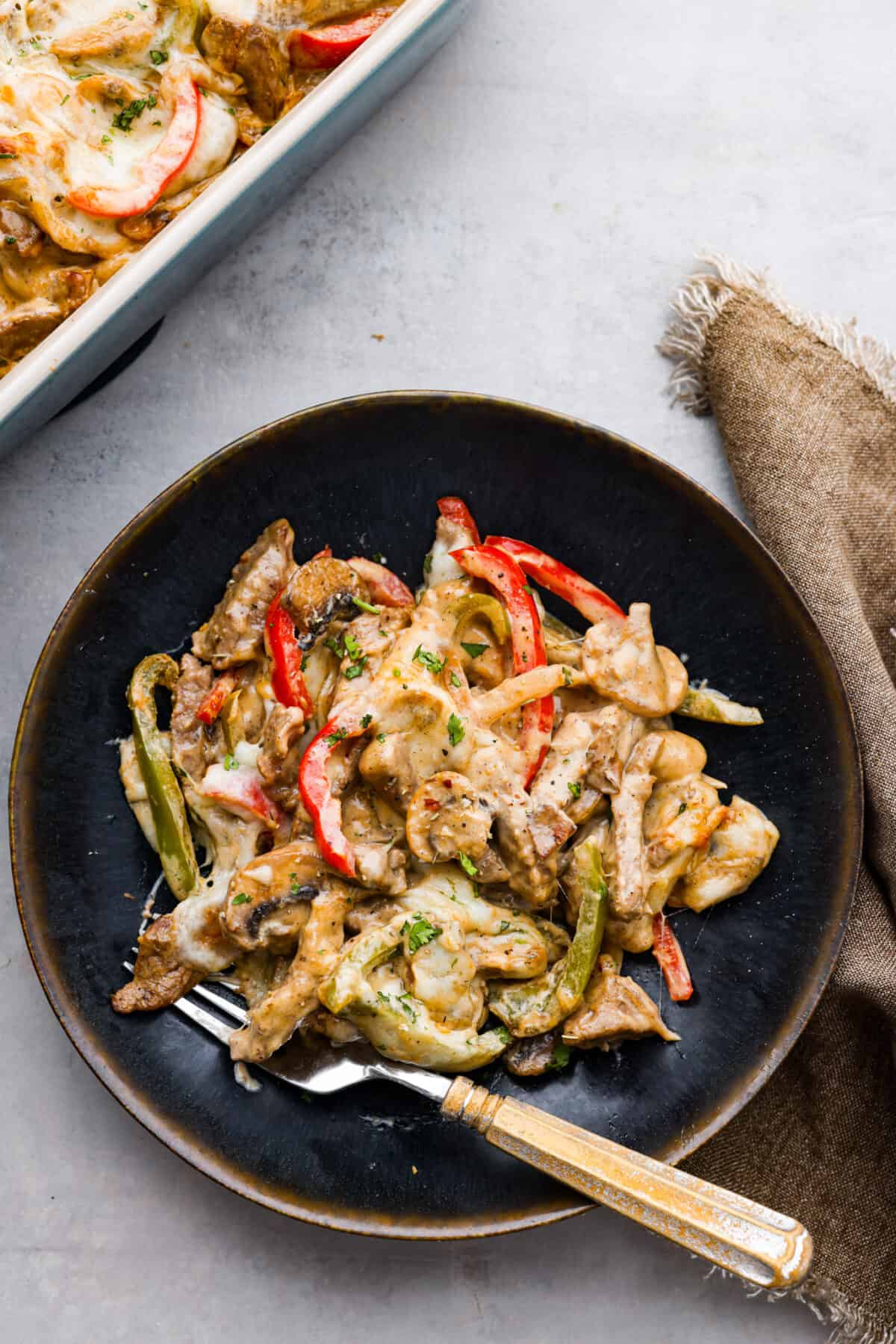 Try this delicious Philly Cheesesteak Casserole, a twist on the classic Philly Cheesesteak. Tender sirloin steak, colorful veggies, and creamy cheese come together for a mouthwatering meal the whole family will love! Philly Cheesesteak Casserole in a black bowl with a fork.