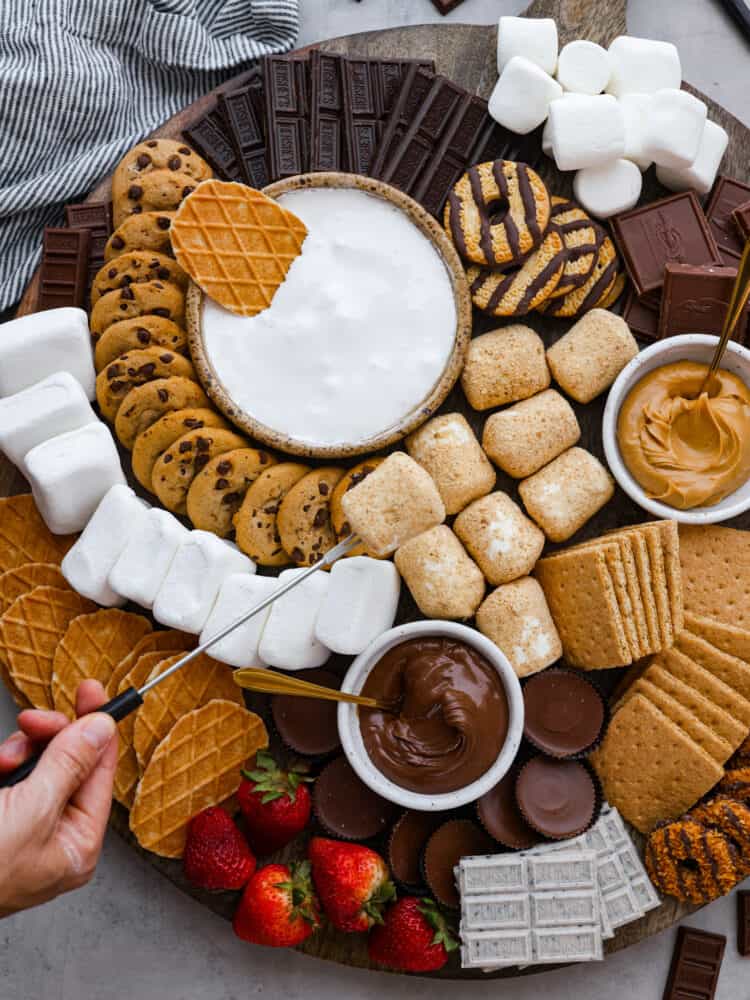 Just in time for summer barbecues and camping trips, this s'mores charcuterie board is here to sweeten things up! It features a mix of different types of chocolate, crackers, marshmallows, and spreads so everyone in your family can create their own perfect sweet treat. Someone holding a skewer with a toasted coconut marshmallow on the end of it.