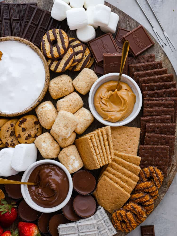 Just in time for summer barbecues and camping trips, this s'mores charcuterie board is here to sweeten things up! It features a mix of different types of chocolate, crackers, marshmallows, and spreads so everyone in your family can create their own perfect sweet treat. Closeup of a sโmores board. In addition to cookies, chocolate, and marshmallows, there are 2 small bowls filled with nutella and peanut butter.