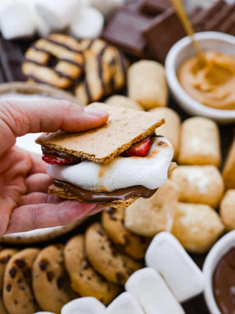 Just in time for summer barbecues and camping trips, this s'mores charcuterie board is here to sweeten things up! It features a mix of different types of chocolate, crackers, marshmallows, and spreads so everyone in your family can create their own perfect sweet treat. A s'more being held is someone's hand.