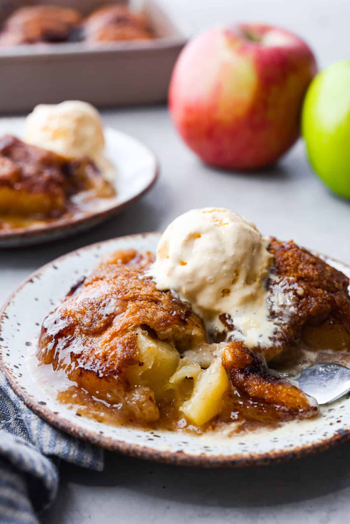 This semi-homemade version of apple dumplings is made with a delicious surprise ingredient! Apple slices get wrapped in crescent dough, smothered in a caramel sauce, then topped with bubbly canned Sprite. You won't believe how incredible this concoction is when it bakes up! Baked apple dumplings on a white speckled plate with a scoop of vanilla ice cream on top.