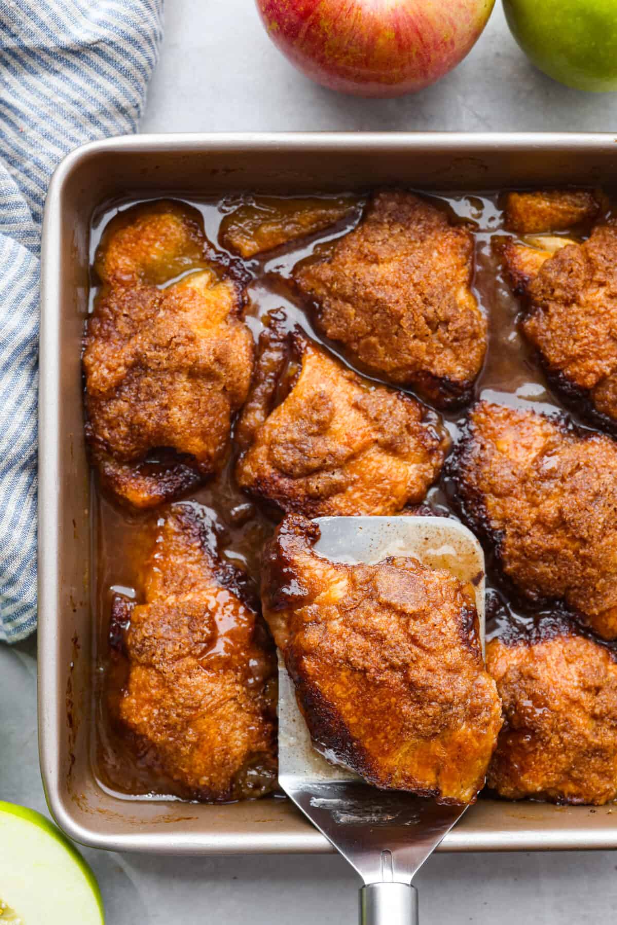 This semi-homemade version of apple dumplings is made with a delicious surprise ingredient! Apple slices get wrapped in crescent dough, smothered in a caramel sauce, then topped with bubbly canned Sprite. You won't believe how incredible this concoction is when it bakes up! Top view of baked apple dumplings in a pan with a spatula. Apples are next to the pan with a striped kitchen towel.