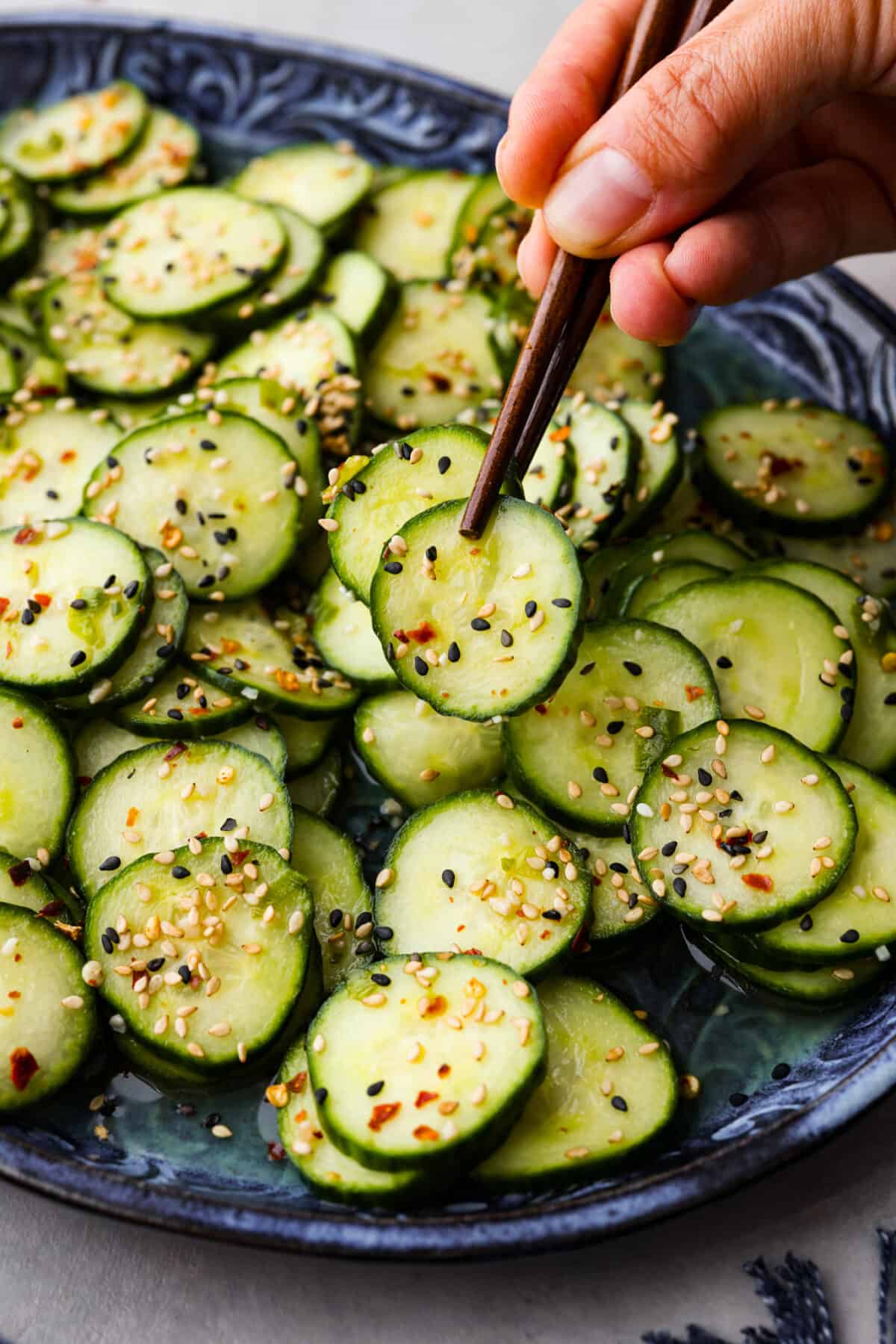 This delightful Asian Cucumber Salad is a blend of flavors creating a refreshing dish that's finished with a sprinkle of red pepper flakes and sesame seeds. It makes the perfect snack or side dish to complement any meal. A pair of chopsticks lifting a slice of cucumber out of the bowl.