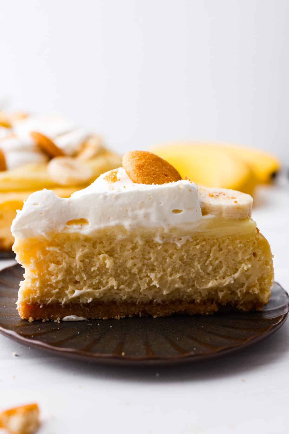 Get ready to enjoy a slice of pure dessert perfection with this Banana Pudding Cheesecake recipe. This yummy dessert has a buttery vanilla wafer crust, a velvety cream cheese filling with ripe bananas, and a classic banana pudding topping. A slice of banana pudding cheesecake on a brown plate.