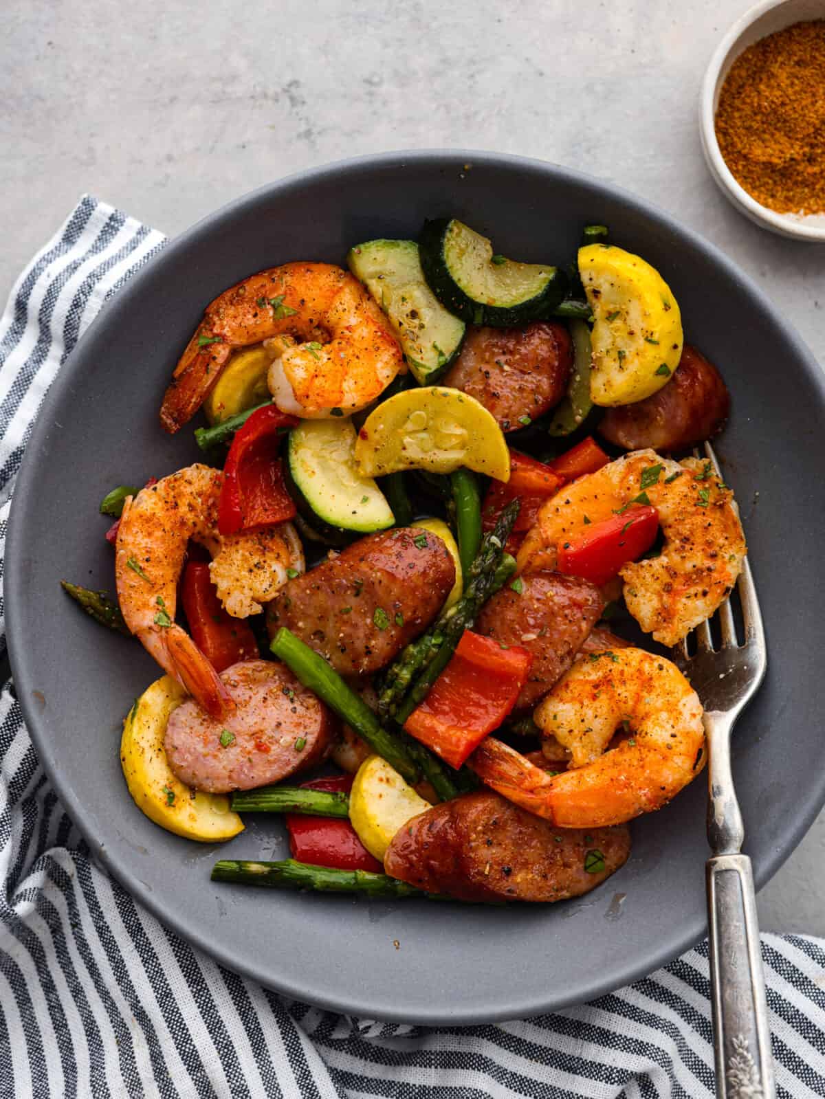 Cajun shrimp and sausage vegetable sheet pan is so incredibly easy that it makes a perfect weeknight meal! It's bursting with amazing Cajun flavor! This is a healthy and delicious one-pan dinner full of shrimp, sausage, and veggies. Close view of Cajun shrimp, sausage, and veggies in a gray bowl with a fork.