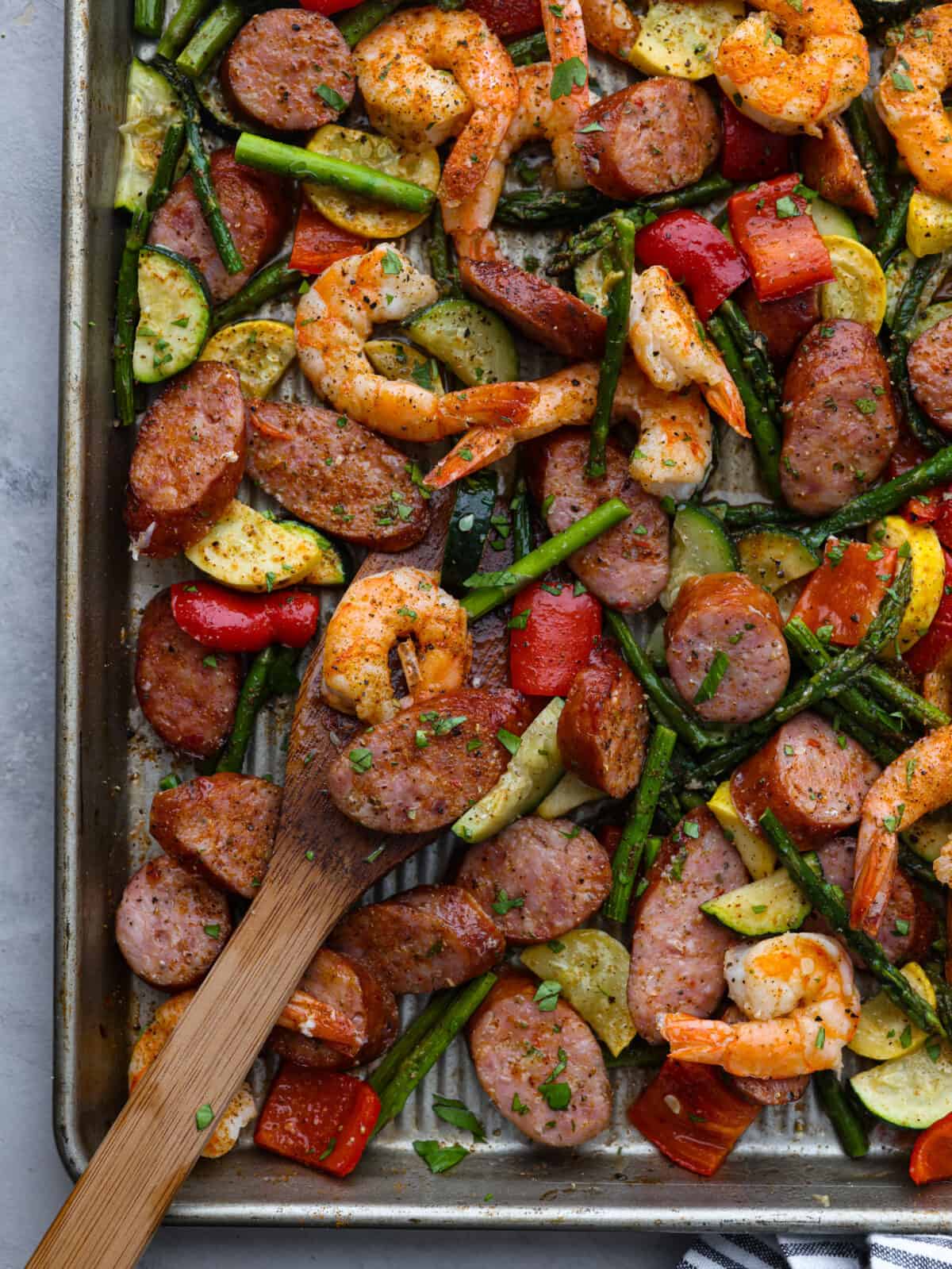 Cajun shrimp and sausage vegetable sheet pan is so incredibly easy that it makes a perfect weeknight meal! It's bursting with amazing Cajun flavor! This is a healthy and delicious one-pan dinner full of shrimp, sausage, and veggies. Close view of cajun shrimp and sausage vegetable sheet pan with a wood serving spoon on the pan.
