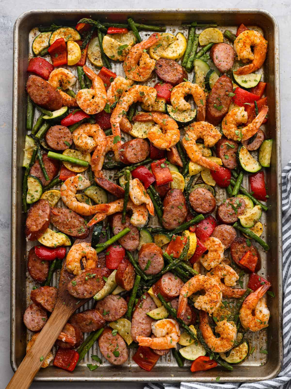 Cajun shrimp and sausage vegetable sheet pan is so incredibly easy that it makes a perfect weeknight meal! It's bursting with amazing Cajun flavor! This is a healthy and delicious one-pan dinner full of shrimp, sausage, and veggies. Top view of cajun shrimp and sausage sheet pan on a sheet pan. A striped towel is next to the sheet pan.
