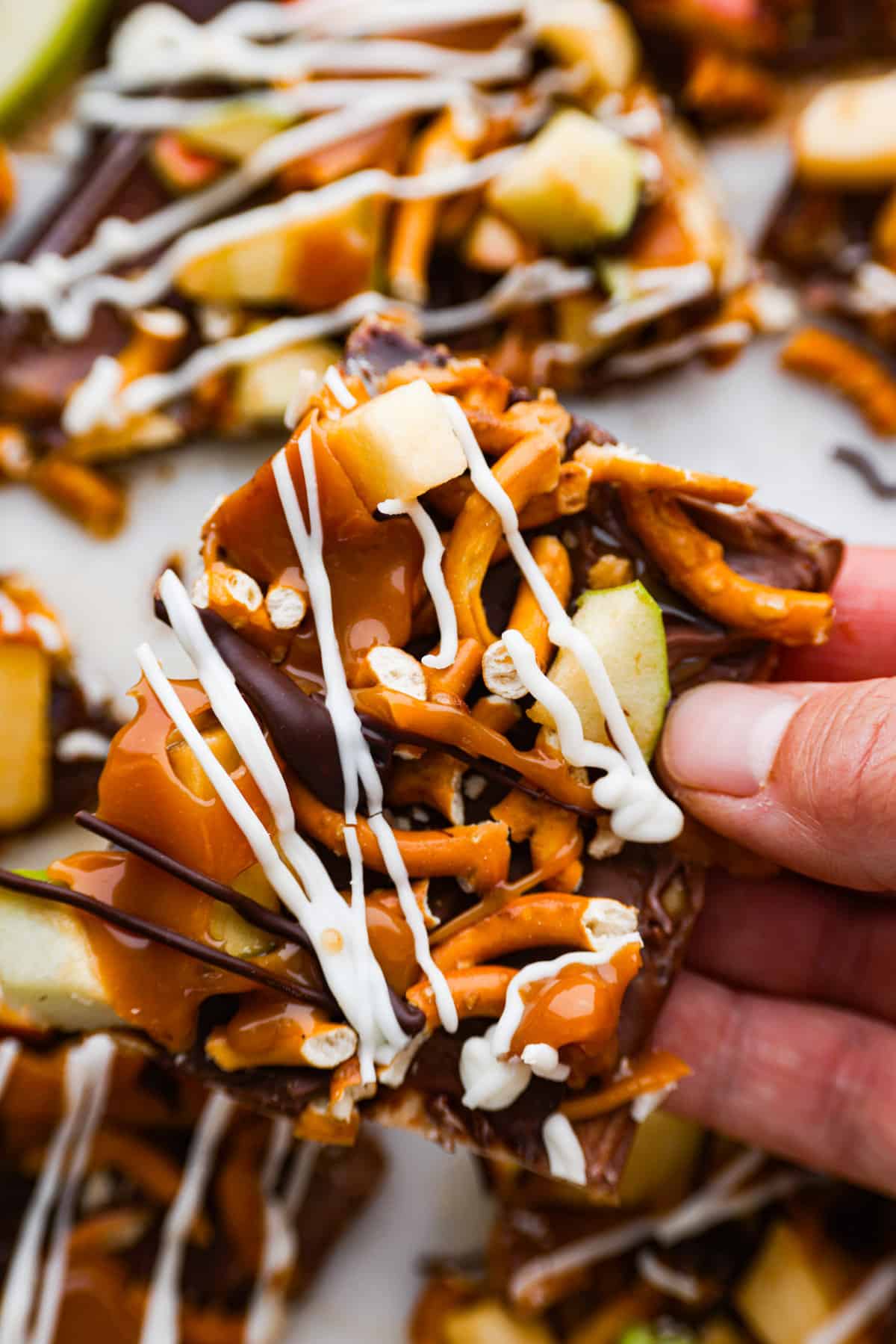 Meet your new favorite fall treat: caramel apple bark made with pretzels and a mix of white and dark chocolate! Itโs rich and sweet with the perfect crunch. One bite and youโll be hooked! Closeup of a piece of bark being held.