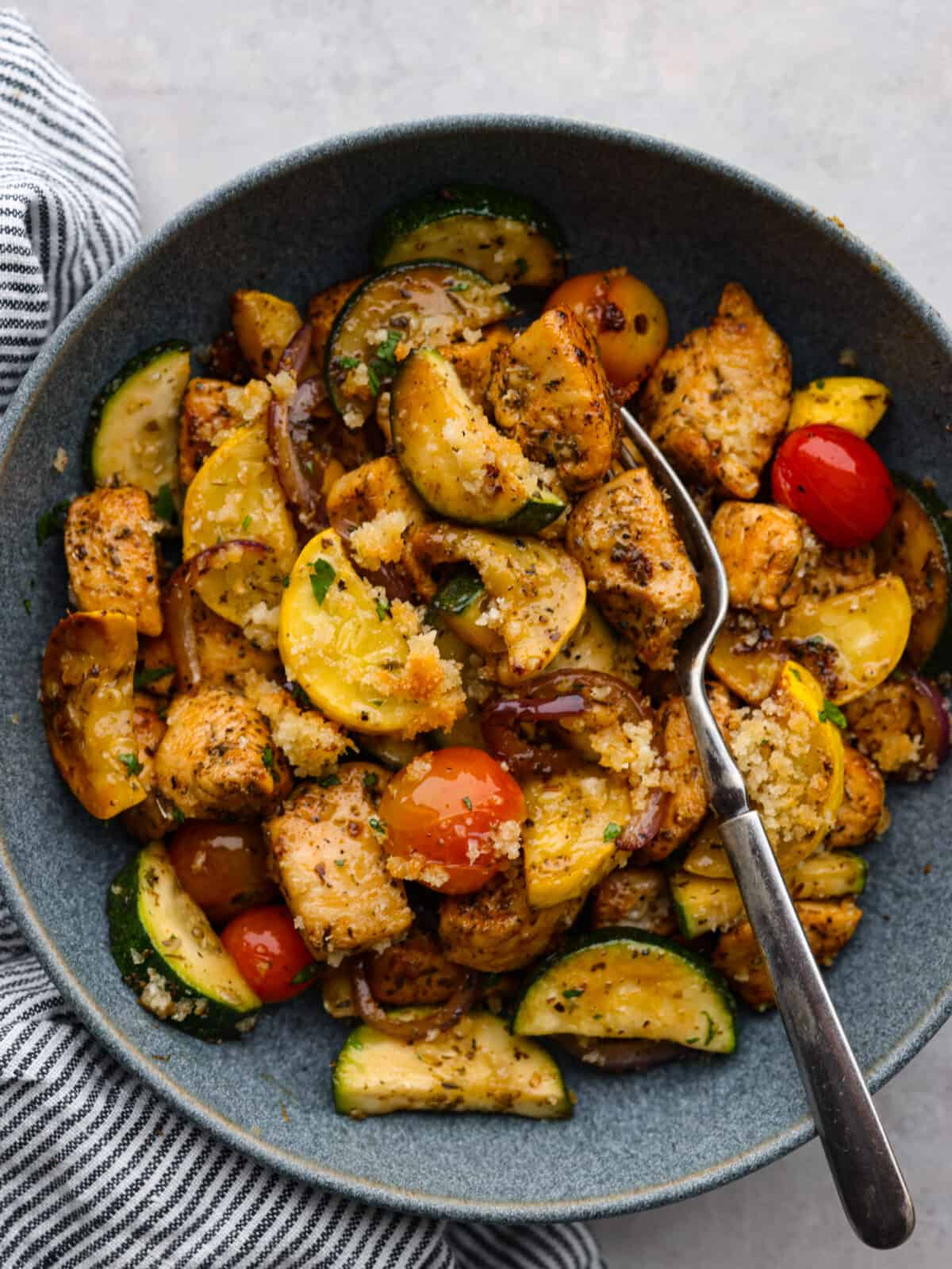 Use up all those garden veggies in this delicious chicken and garden vegetable skillet. Your family will love this healthy 30-minute skillet dinner full of seasoned chicken, sauteed vegetables, and a cheesy breadcrumb topping. Close view of chicken and garden vegetables in a gray bowl with a fork in the bowl.