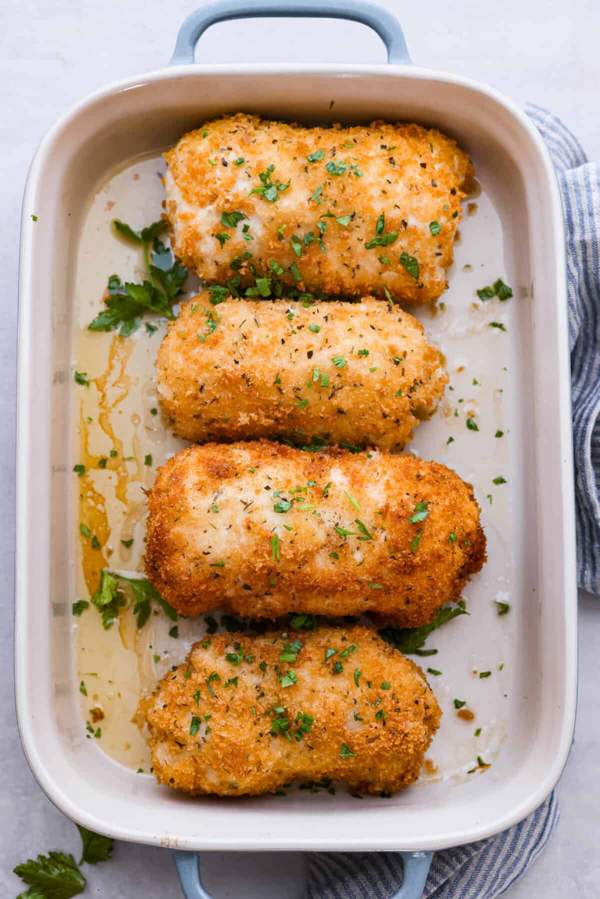 Level up dinnertime with this crispy Chicken Kiev! Each piece of chicken is filled with herby compound butter, then coated in bread crumbs and fried to create a golden crust. Crispy on the outside, extra moist and flavorful on the inside, this is the perfect dinner for any occasion. Top-down view of the chicken in a blue and white casserole dish.