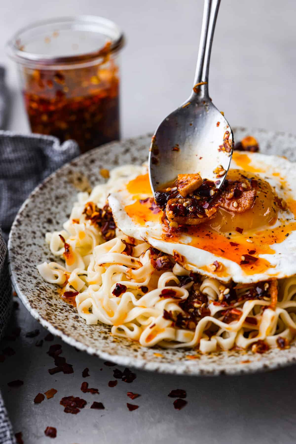 Chili crisp is the secret ingredient every kitchen needs to spice up your dishes effortlessly. It's a mix of crispy shallots, garlic, ginger, fiery chili flakes and sesame seeds that will add the best flavor to any dish! Chili oil being added to an egg and noodle dish.