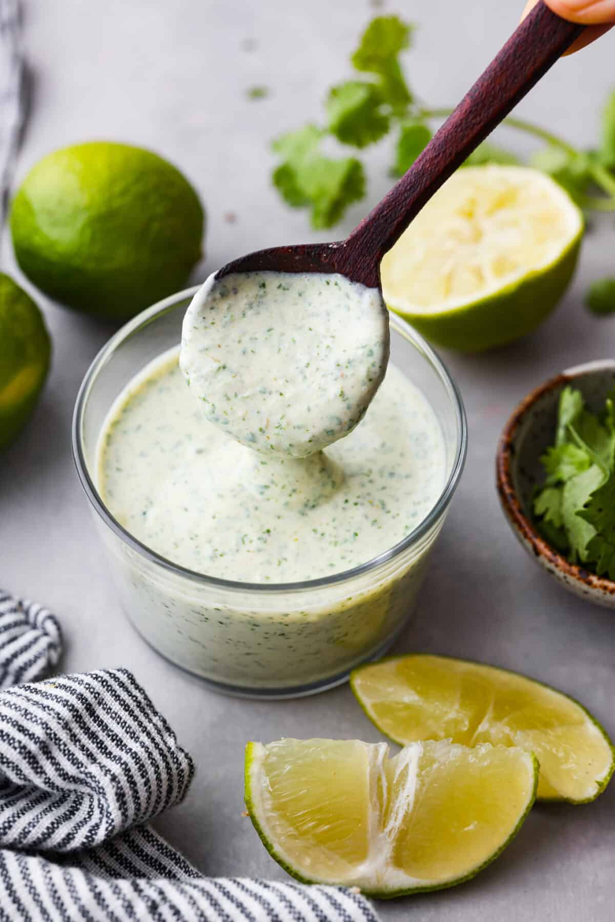 Once you taste this creamy cilantro lime sauce, you'll want to drizzle it on everything in sight! It's a tasty mix of sour cream, cilantro, salsa verde, lime juice, garlic, salt, and pepper. Perfect for grilled meats, salads, tacos, and more! Hero image of cilantro lime sauce in a glass jar, being mixed with a wooden spoon.