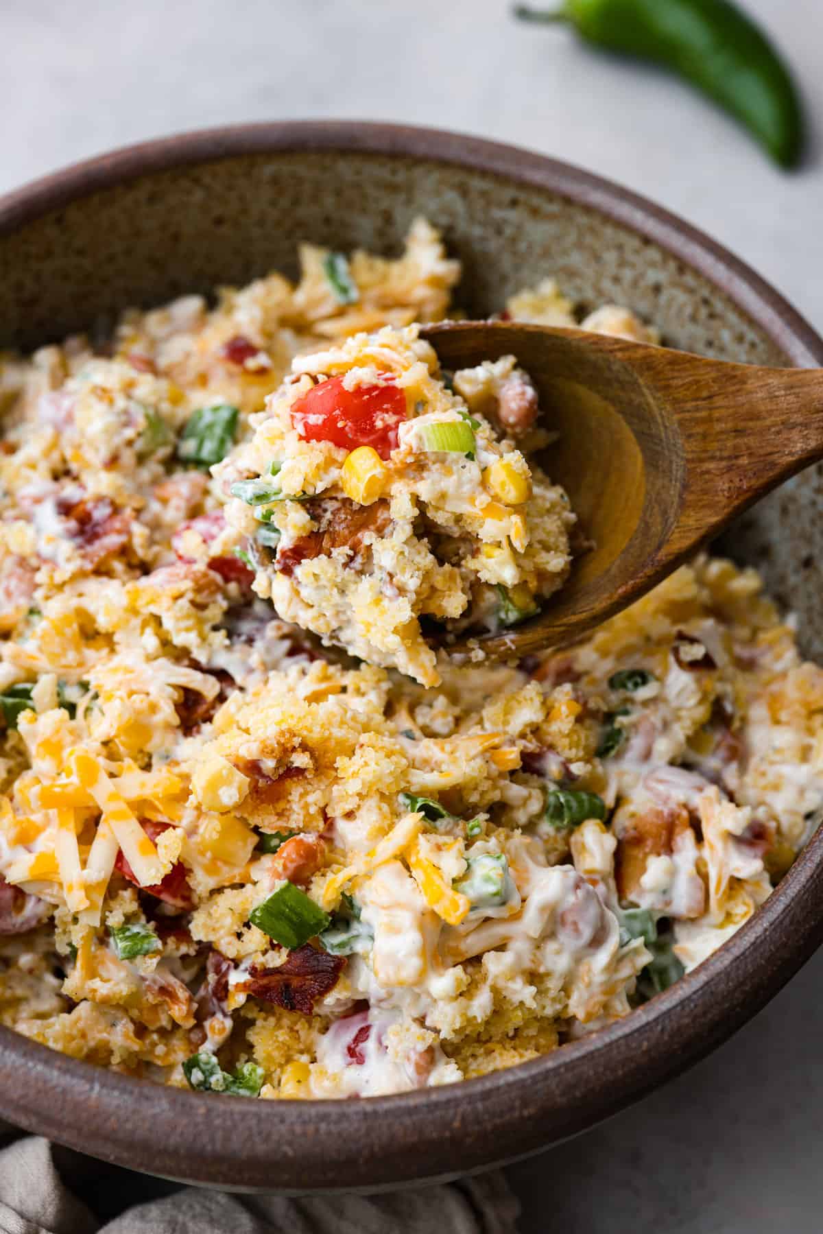 Tasty cornbread salad is a classic Southern salad bursting with comforting flavors. Sweet buttery cornbread gets tossed together with tomatoes, corn, pinto beans, bacon crumbles, green onions, and shredded cheese. A creamy ranch dressing perfectly marries these delicious salad ingredients together. Close view of cornbread salad in a bowl with a wood spoon lifting up a spoonful of salad.
