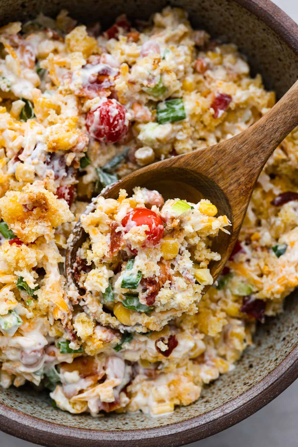 Tasty cornbread salad is a classic Southern salad bursting with comforting flavors. Sweet buttery cornbread gets tossed together with tomatoes, corn, pinto beans, bacon crumbles, green onions, and shredded cheese. A creamy ranch dressing perfectly marries these delicious salad ingredients together. Close view of cornbread salad in a bowl with a wood spoon lifting up a spoonful of salad.