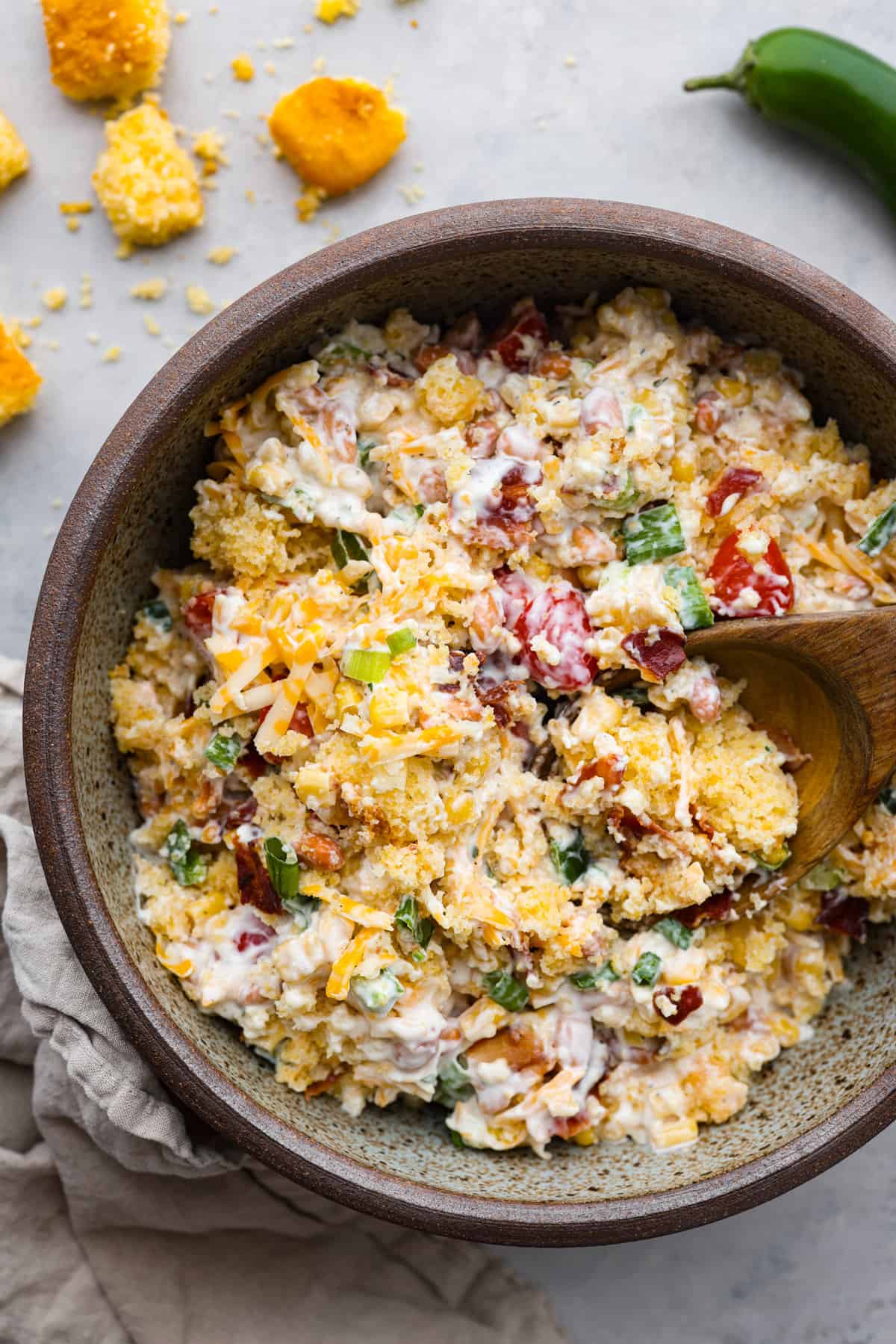 Tasty cornbread salad is a classic Southern salad bursting with comforting flavors. Sweet buttery cornbread gets tossed together with tomatoes, corn, pinto beans, bacon crumbles, green onions, and shredded cheese. A creamy ranch dressing perfectly marries these delicious salad ingredients together. Top view of cornbread salad in a large brown bowl with a wooden spoon inside the bowl.