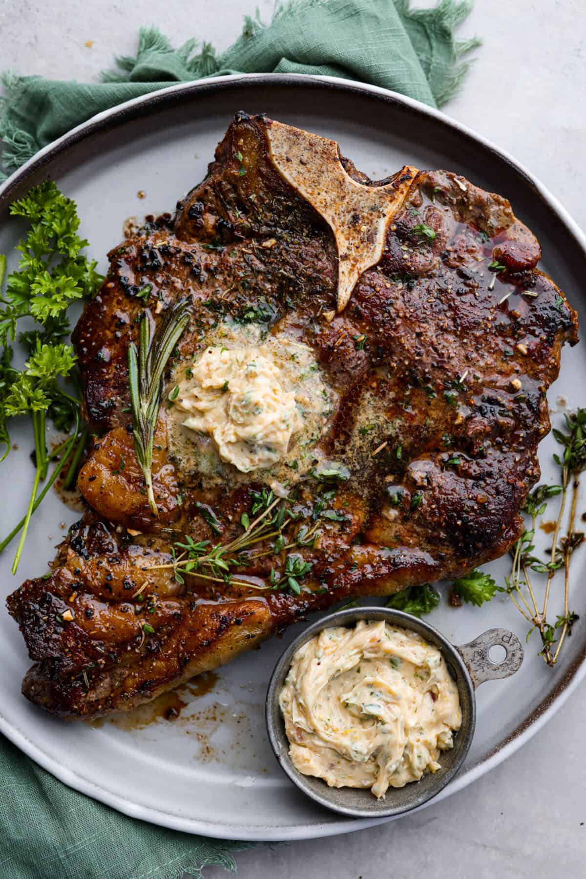 Anything smothered with this flavor-packed cowboy butter will satisfy your tastebuds! Soft butter mixed with garlic, shallot, fresh herbs, and seasonings that take your butter up a notch. Enjoy on top of meat, slathered on bread, or melted on a baked potato. Top view of cowboy butter melted on top of a steak. A bowl of cowboy butter is next to the steak on a gray plate.