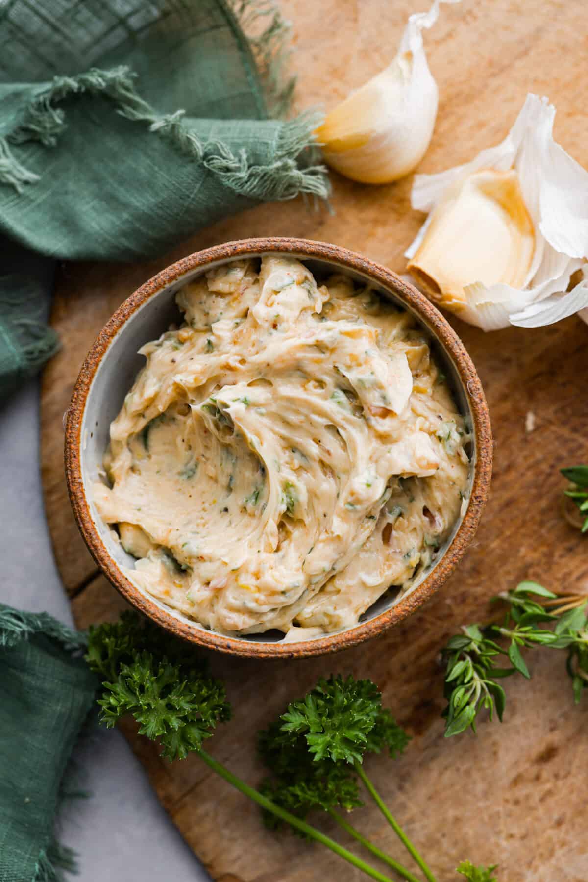 Anything smothered with this flavor-packed cowboy butter will satisfy your tastebuds! Soft butter mixed with garlic, shallot, fresh herbs, and seasonings that take your butter up a notch. Enjoy on top of meat, slathered on bread, or melted on a baked potato. Top view of cowboy butter in a rustic bowl on a wood board with herbs and garlic next to the bowl.