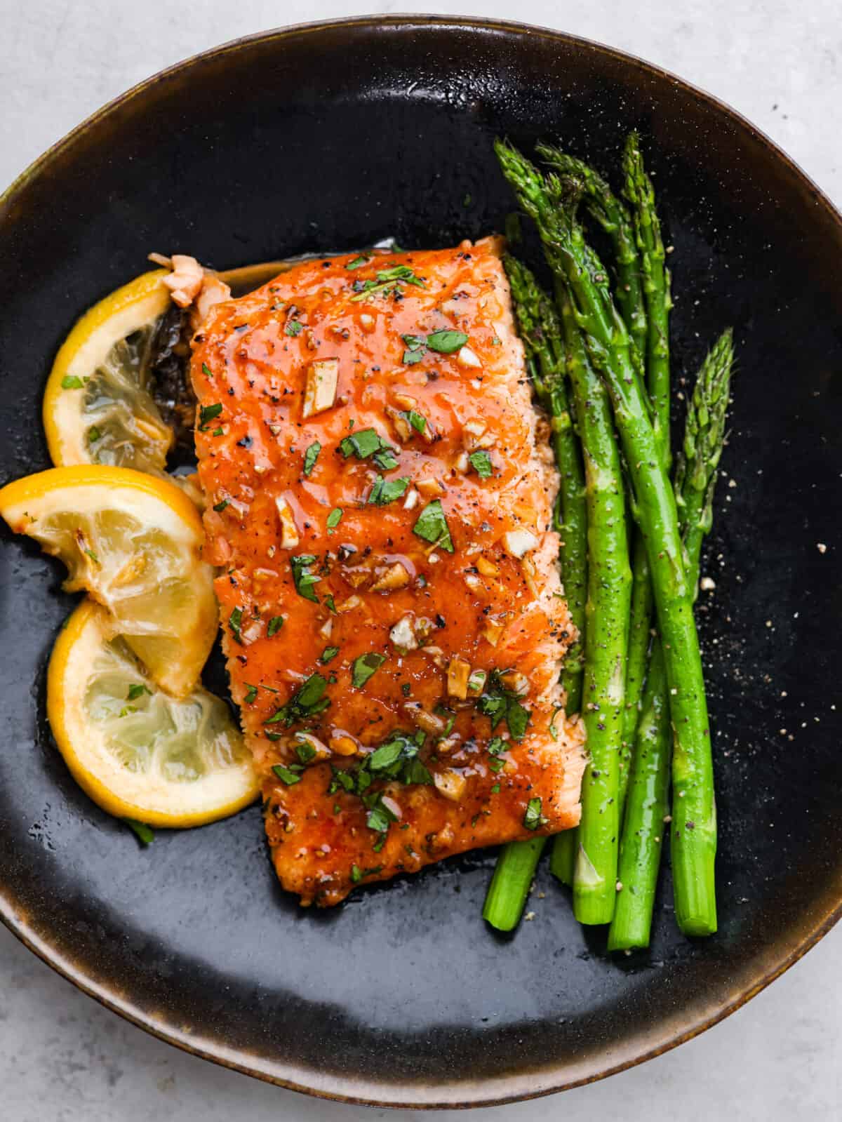 Garlic Brown Sugar Glazed Salmon is tender and flaky salmon covered in the most amazing garlic brown sugar soy glaze. The flavor is out of this world, and it will become a new family favorite! A black plate with garlic brown sugar salmon, lemon slices and asparagus.