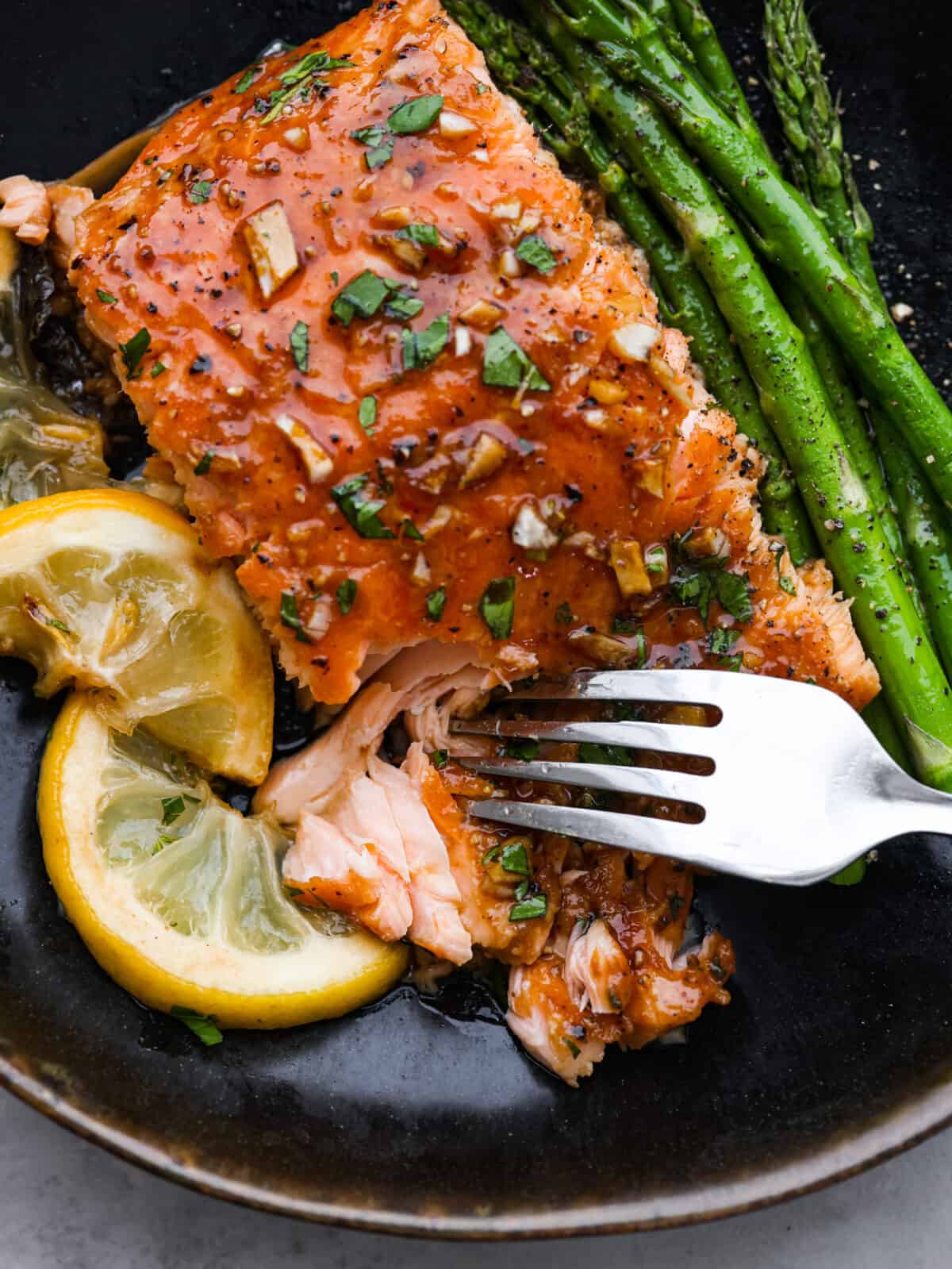 Garlic Brown Sugar Glazed Salmon is tender and flaky salmon covered in the most amazing garlic brown sugar soy glaze. The flavor is out of this world, and it will become a new family favorite!ย A close up of garlic salmon on a plate with asparagus and lemon slices being cut into with a fork.