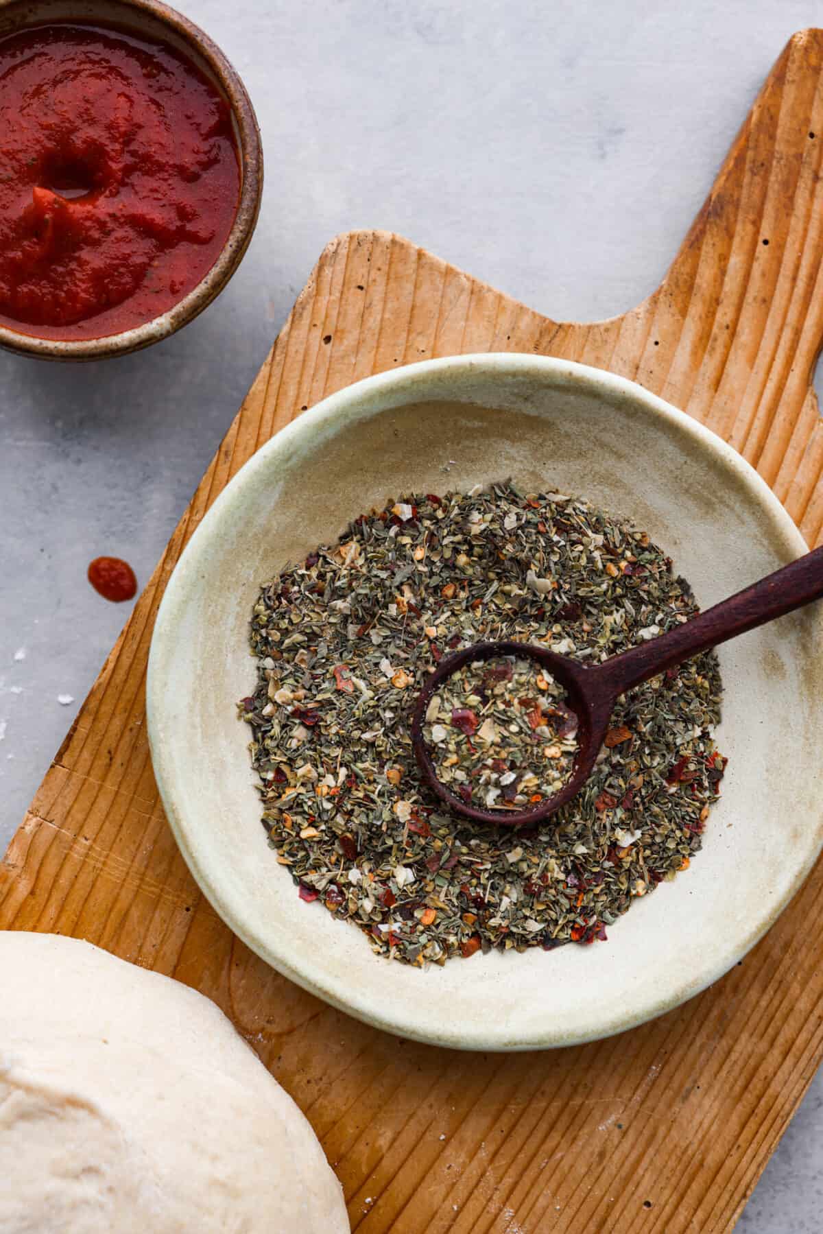 Add this homemade pizza seasoning to all of your favorite pizzas and watch them transform! It's the best blend of herbs, garlic, and red pepper flakes to make every bite a dream! A small bowl of pizza seasoning on a wooden board.