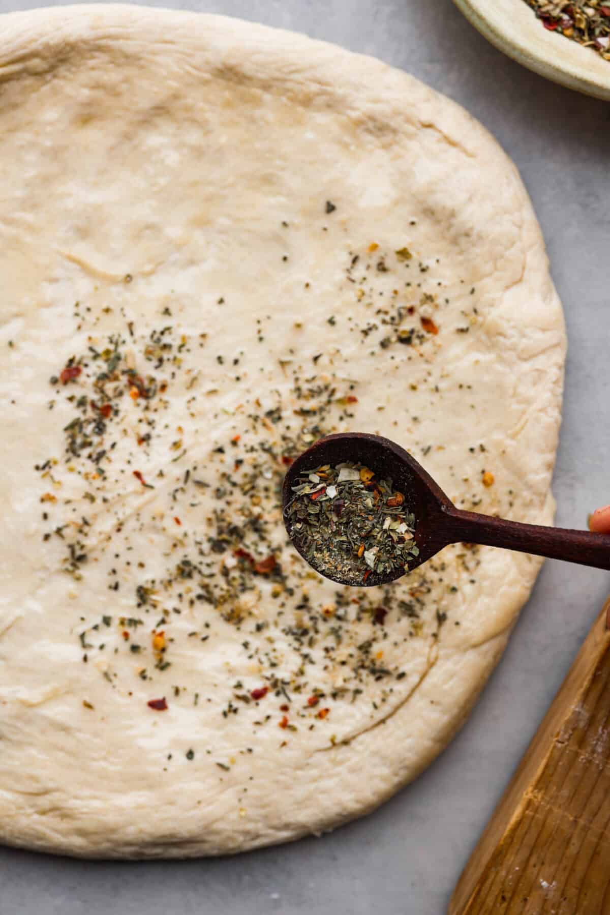 Add this homemade pizza seasoning to all of your favorite pizzas and watch them transform! It's the best blend of herbs, garlic, and red pepper flakes to make every bite a dream! Pizza seasoning being sprinkled over dough.