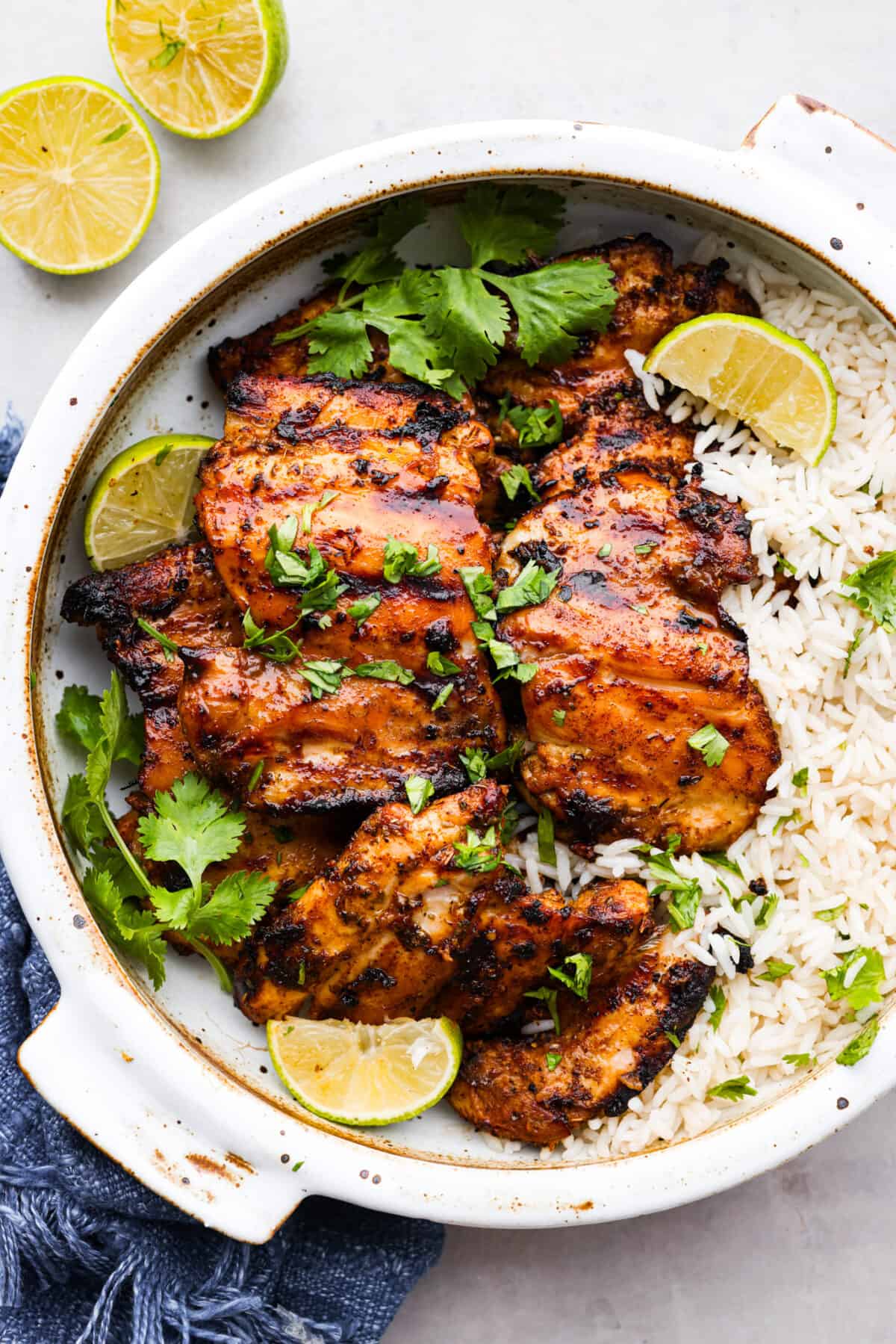 Mexican pollo asado is some of the juiciest, most flavorful grilled chicken you'll ever taste. Each chicken thigh is marinated in a mix of tangy lime, orange, and a blend of spices for some kick. You'll love each tender, flavor-packed bite! Mexican grilled chicken served over rice. It's garnished with fresh herbs and lime slices.