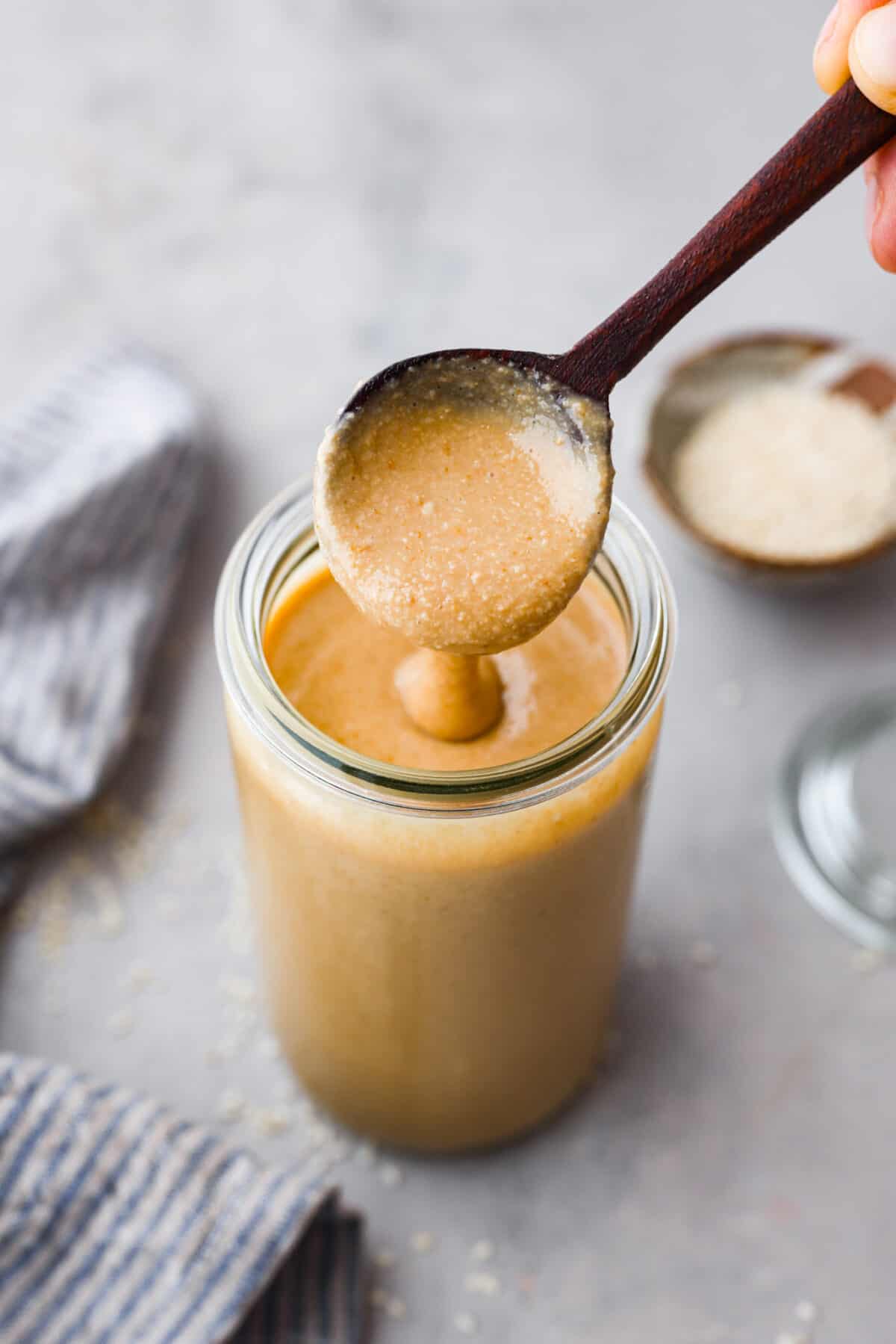 Make this easy tahini right at home with just sesame seeds and olive oil. This Middle Eastern condiment resembles the same texture as nut butter and is a staple ingredient used in sauces, savory dishes, and even desserts! Top view of tahini in a glass jar with a wood spoon lifting up spoonful of tahini.