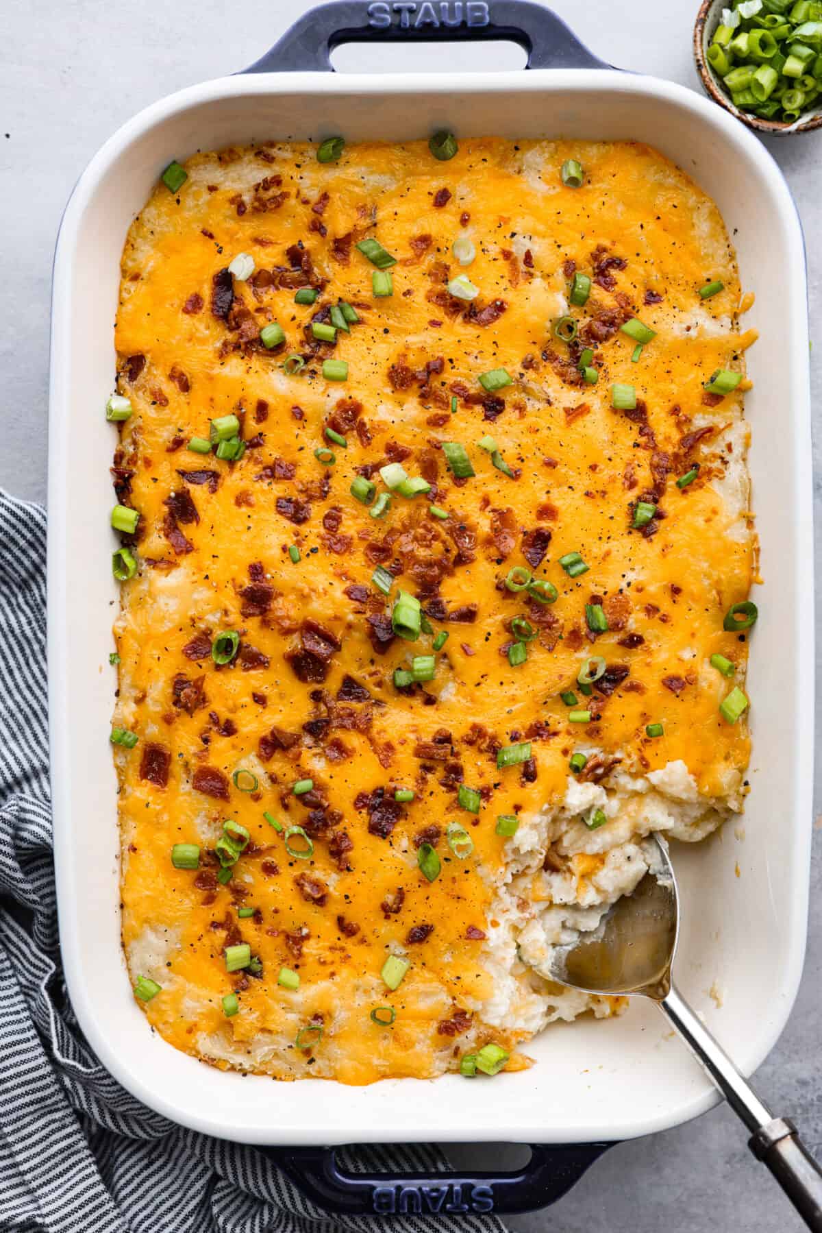 Introducing the BEST twice baked potato casserole you will ever make! Loaded with tender baked potatoes, bacon, and cheese then baked together in a delicious creamy casserole. Top view of twice baked potato casserole in a blue casserole dish and garnished with green onions.