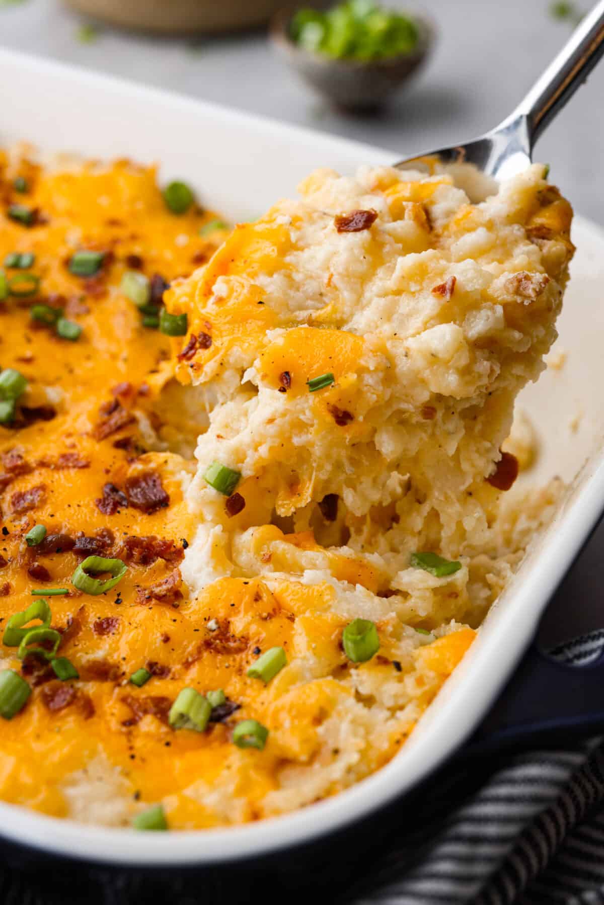 Introducing the BEST twice baked potato casserole you will ever make! Loaded with tender baked potatoes, bacon, and cheese then baked together in a delicious creamy casserole. Close side view of twice baked potato casserole lifting out of a casserole dish.