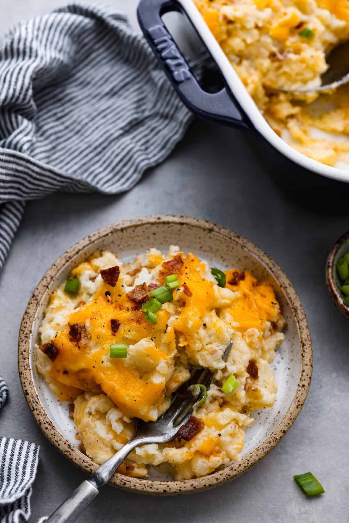 Introducing the BEST twice baked potato casserole you will ever make! Loaded with tender baked potatoes, bacon, and cheese then baked together in a delicious creamy casserole. Top close view of a small plate of twice baked potato casserole. A fork is on the plate and the casserole dish next to it.