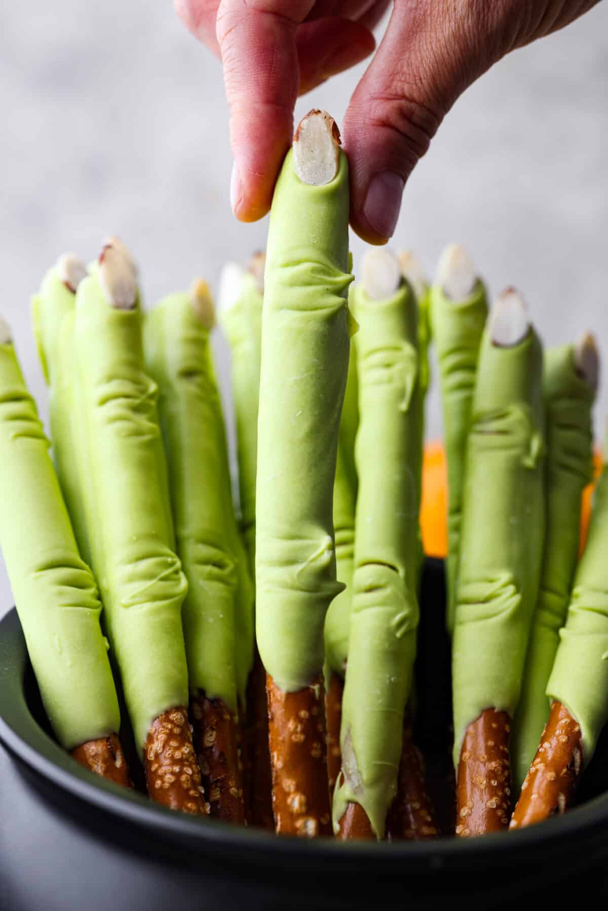 Get ready for a spooky Halloween delight with these Witch Fingers! A classic twist on pretzel rods, this bewitching treat is dipped in green candy melts and decorated with almond nails and knuckles. It makes for a fun and easy treat that's perfect for the season. A close up of witch fingers in a cauldron and a hand pulling one of them out