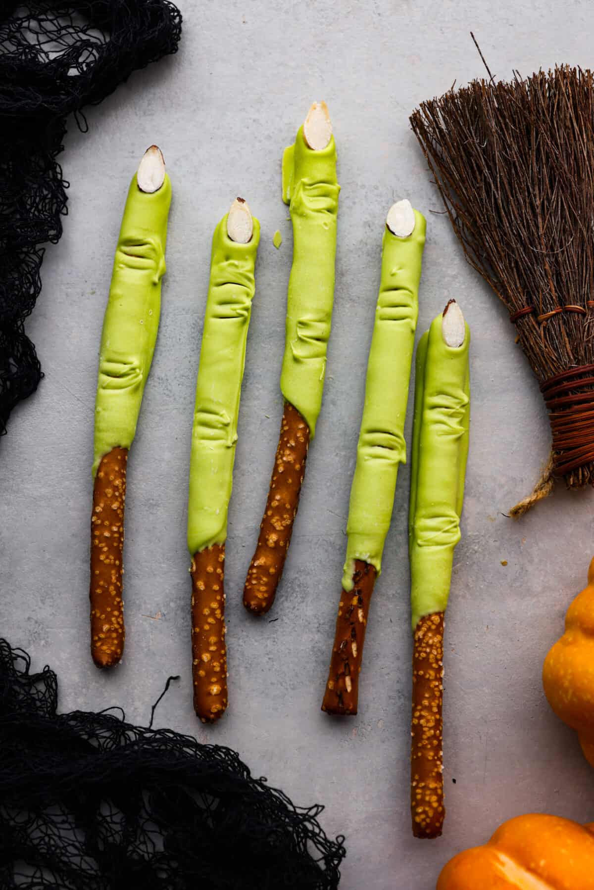 Get ready for a spooky Halloween delight with these Witch Fingers! A classic twist on pretzel rods, this bewitching treat is dipped in green candy melts and decorated with almond nails and knuckles. It makes for a fun and easy treat that's perfect for the season. 5 witch fingers laying down next to each other.