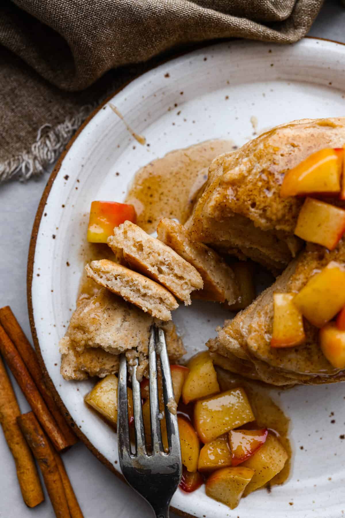 Fluffy applesauce pancakes are easy and delicious with a hint of brown sugar and cinnamon for a cozy fall flavor. The addition of applesauce gives these homemade pancakes the perfect texture. These will quickly become your favorite fall breakfast! Top view of a fork cutting into applesauce pancakes. Sauteed apples and syrup are on the plate of pancakes.