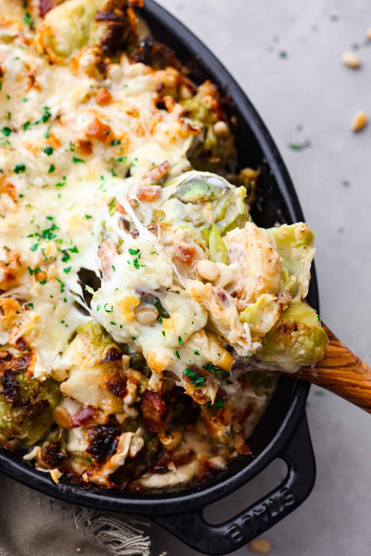 Cheesy and savory Brussels Sprout Casserole is easy to make and perfect for family dinners or holiday meals! Tender, crisp brussels sprouts are combined with pine nuts, bacon and cheese and baked until tender and golden brown. This side dish pairs with a variety of mains and is sure to be a hit! A scoop of casserole on a wooden spoon.