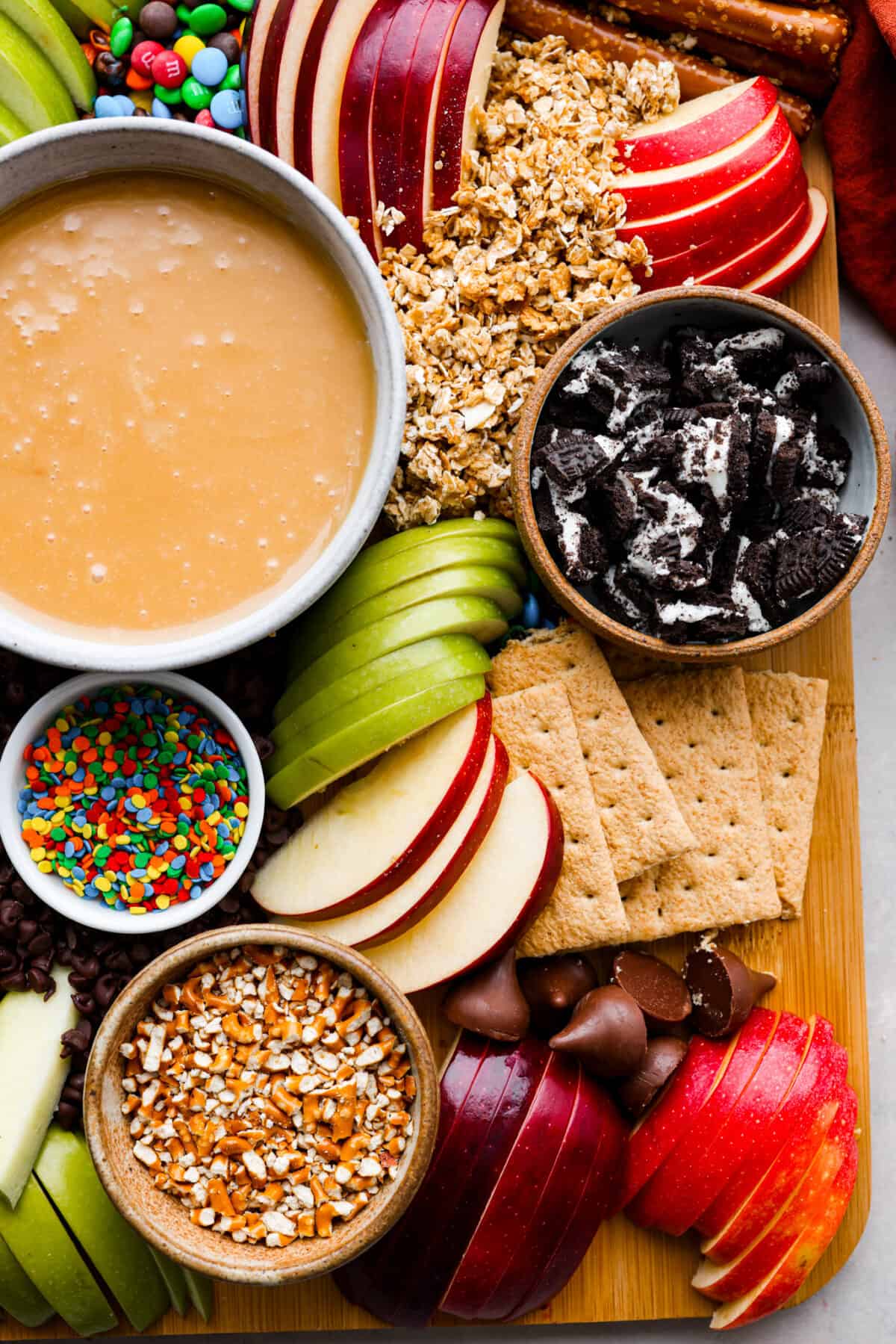 Everybody loves a good caramel apple, especially when they can customize it just the way they like! Make this caramel apple charcuterie board for your next fall party, I promise it will be a huge hit! It has a fun variety of toppings and things for dipping, so thereโs something for everyone! Closeup of caramel sauce in a white bowl as well as oreos, apple slices, m&ms, and granola.