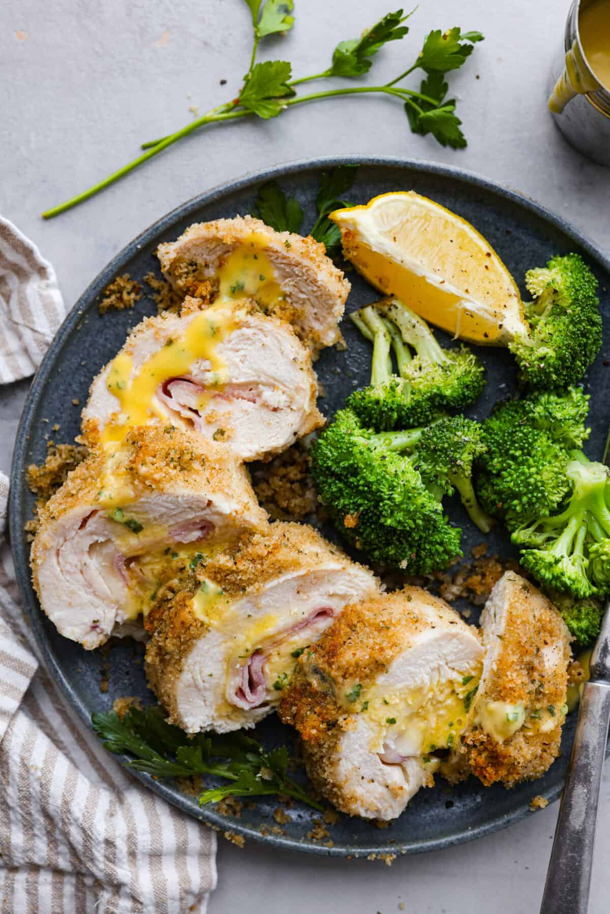 Chicken cordon bleu has a delicious layer of ham and cheese wrapped inside succulent crumb-coated chicken breast. Drizzled with mustard cream sauce, this oven-baked chicken is a classic French recipe your family will love! Overhead shot of plated chicken cordon bleu drizzled with sauce and broccoli and a lemon wedge on the side.