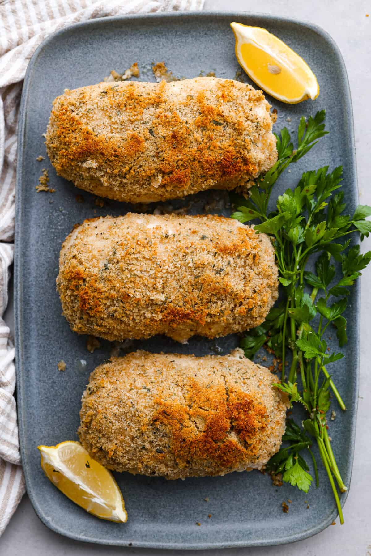 Chicken cordon bleu has a delicious layer of ham and cheese wrapped inside succulent crumb-coated chicken breast. Drizzled with mustard cream sauce, this oven-baked chicken is a classic French recipe your family will love! Top view of chicken cordon bleu on a gray platter garnished with lemon slices and fresh parsley leaves.