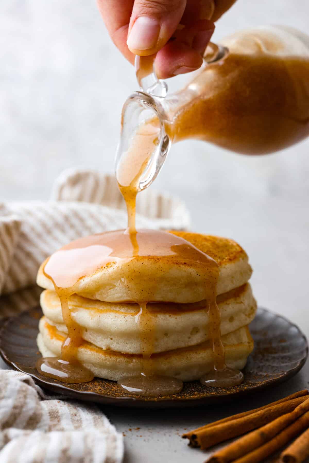 Cinnamon Syrup is the best way to take your pancakes to the next level! It's creamy and buttery and adds delicious cinnamon roll flavor to pancakes, waffles, or anything else you want to sweeten up! Side shot of someone pouring cinnamon syrup over a stack of pancakes.