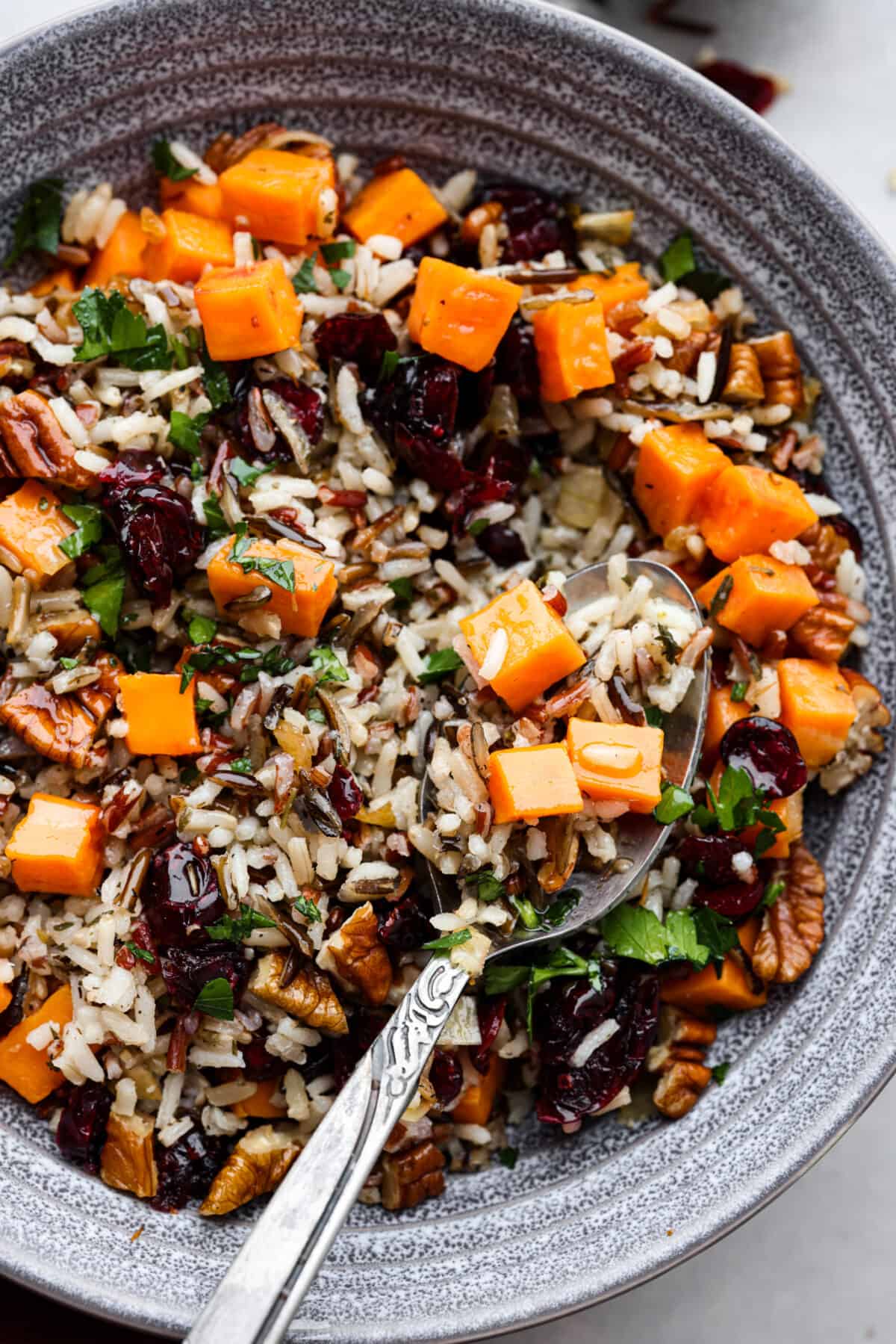 Cranberry pecan sweet potato wild rice pilaf is a perfect fall side dish infused with incredible flavors and textures. Crunchy pecans, cranberries, sweet potato, wild rice, and herbs come together in this tasty dish! Close top view of cranberry pecan sweet potato wild rice pilaf in a gray bowl with a spoon. The spoon is lifting up the pilaf.